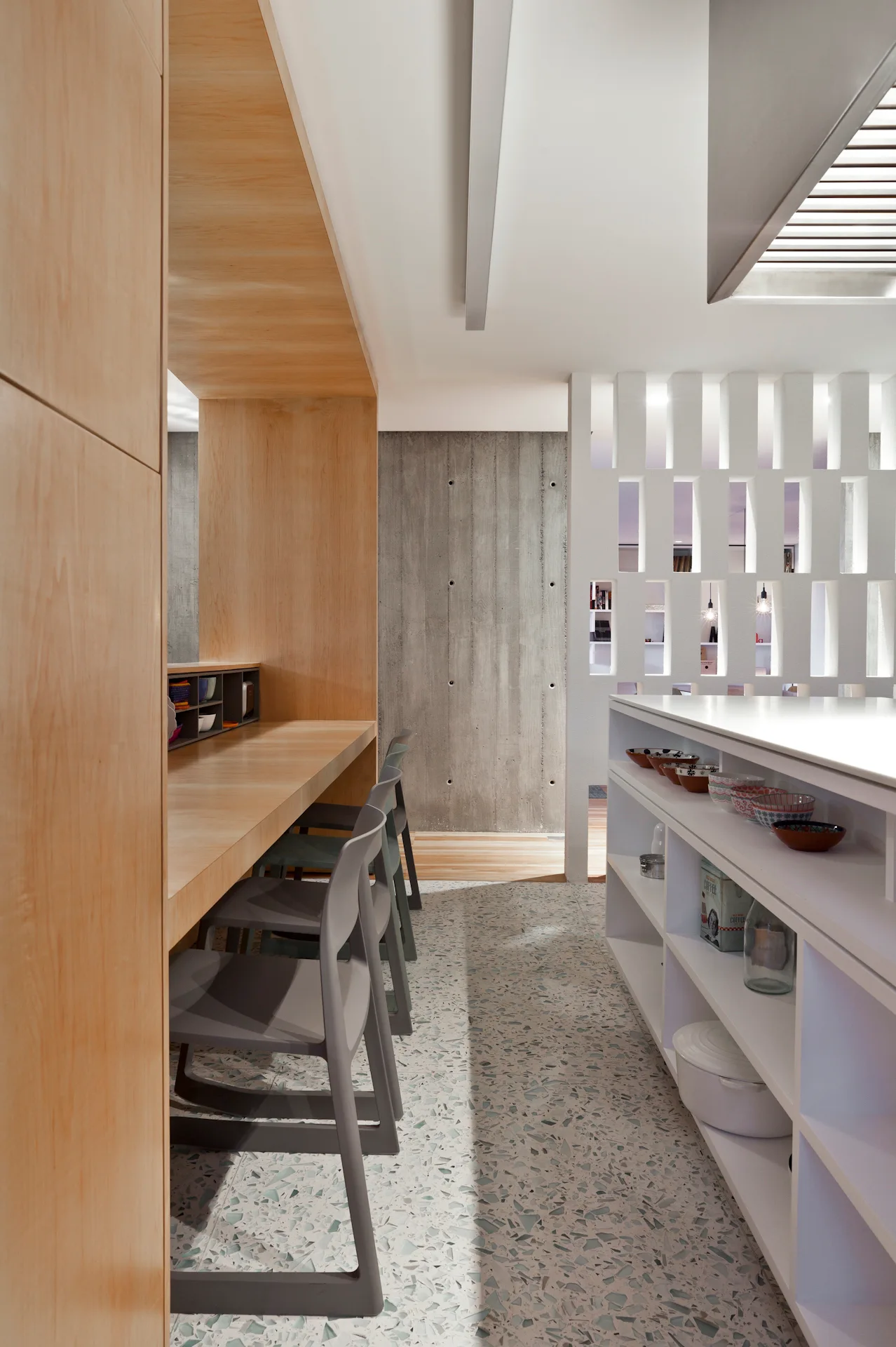 A modern kitchen features a built-in wooden breakfast bar with gray chairs, a white storage island, and speckled terrazzo flooring.