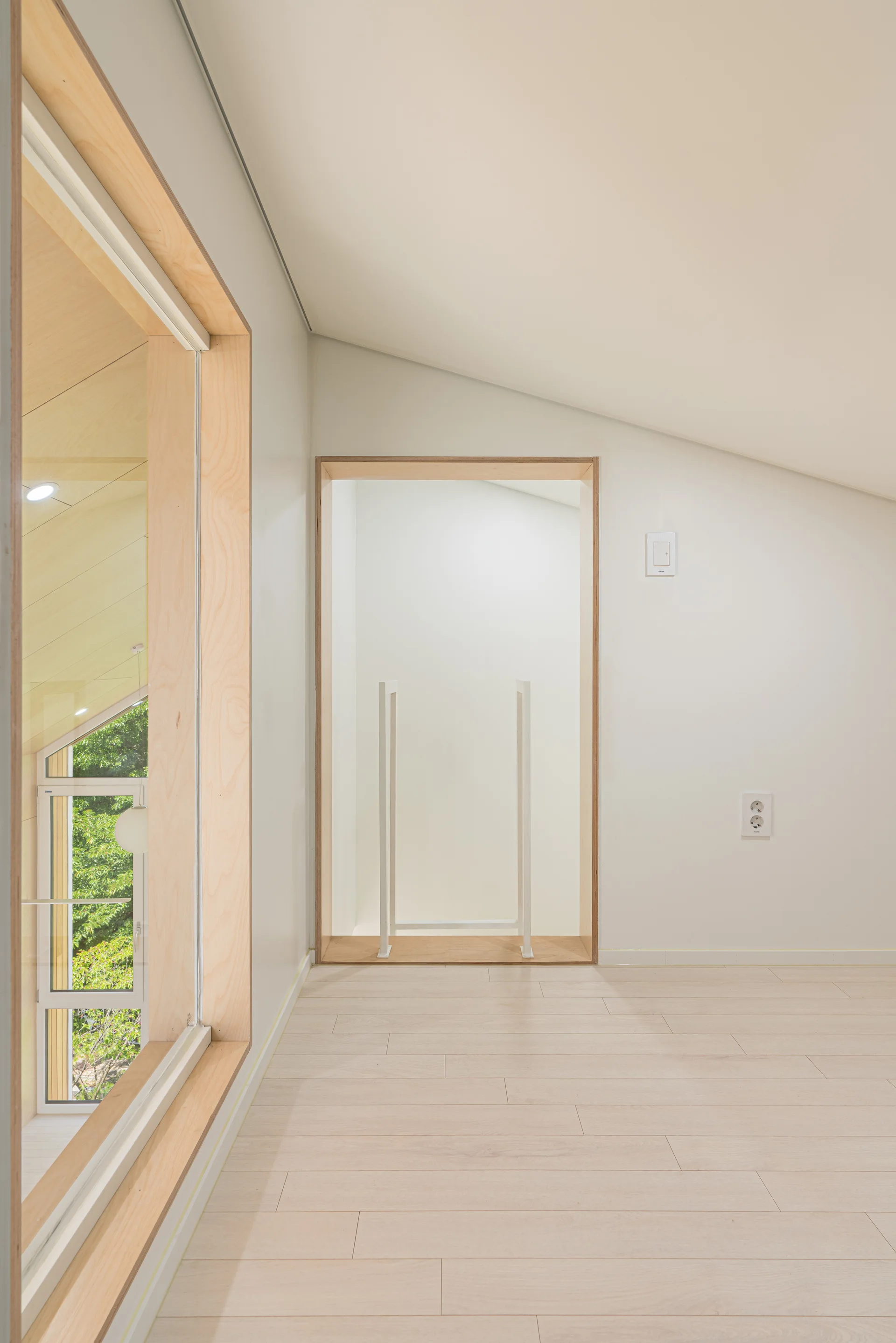 An airy minimalist loft features light wood laminate floors, a sloped ceiling, and large openings framed in thick, pale wood trim.