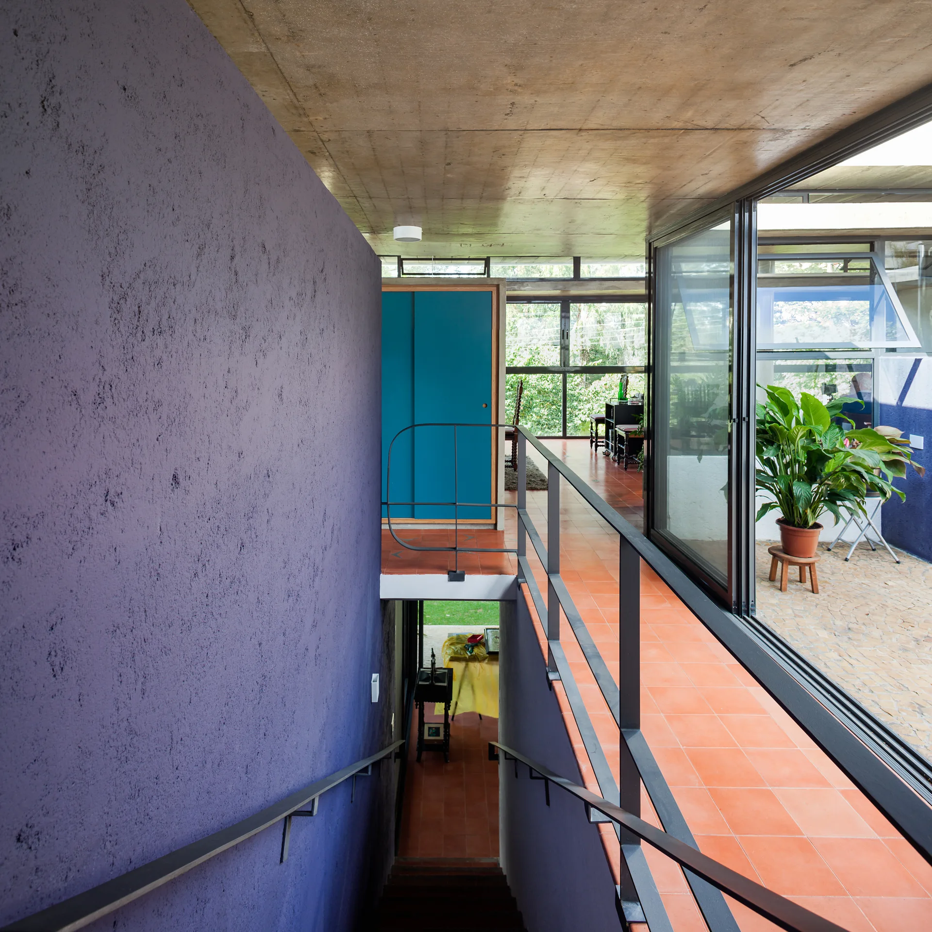 A modern split-level hallway features a staircase beside a textured purple wall, a walkway with terra cotta floor tiles, and an exposed concrete ceiling.
