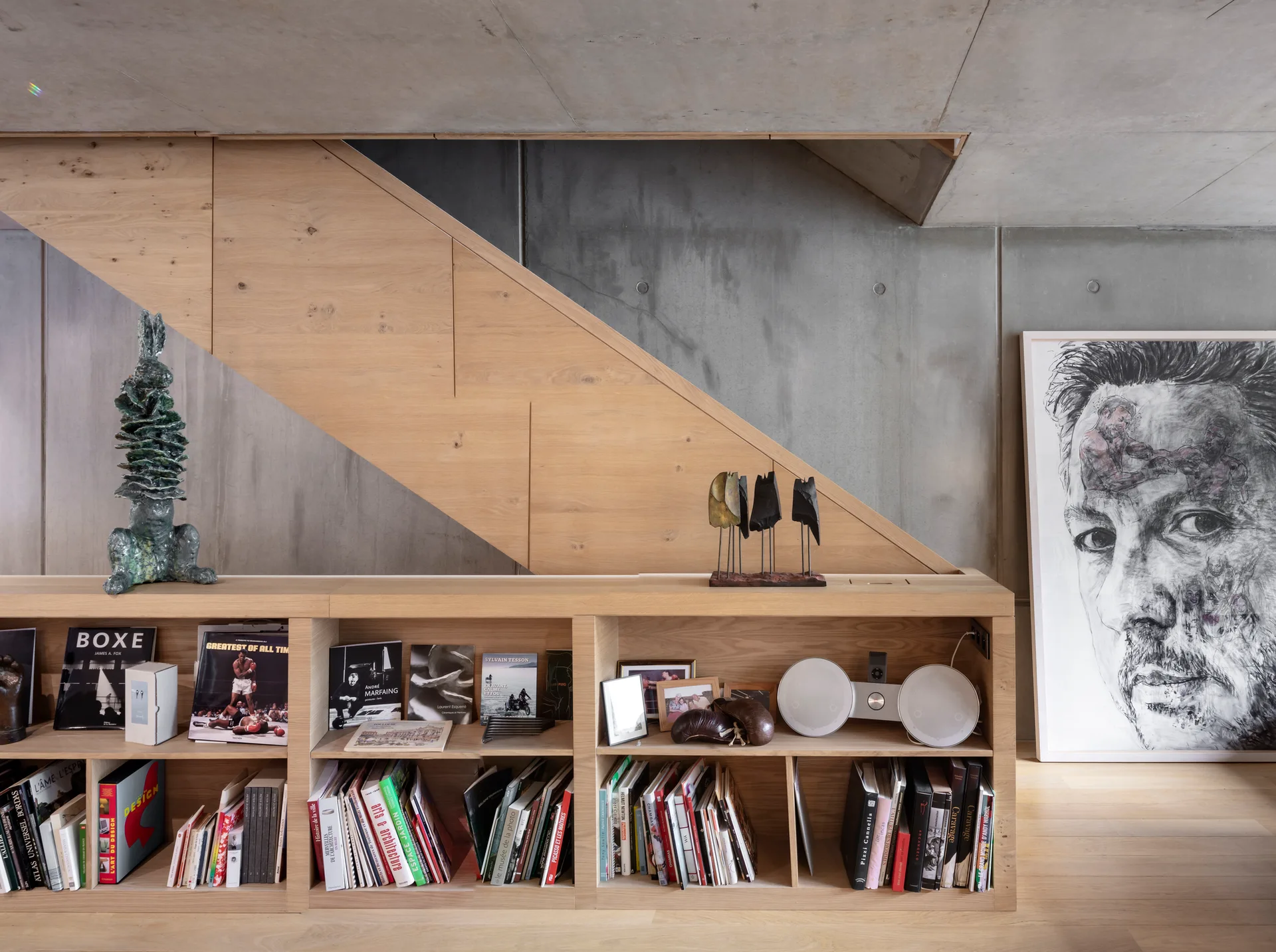 A low-profile light wood bookshelf stands against an industrial concrete wall beneath a matching wood staircase with a large framed portrait on the floor.