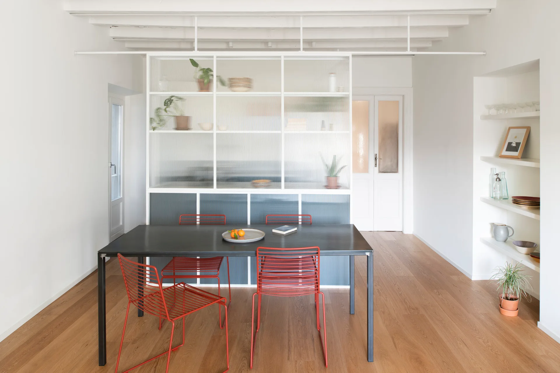 A minimalist dining room features a black table, red wire chairs, and a fluted glass partition under a white beamed ceiling.