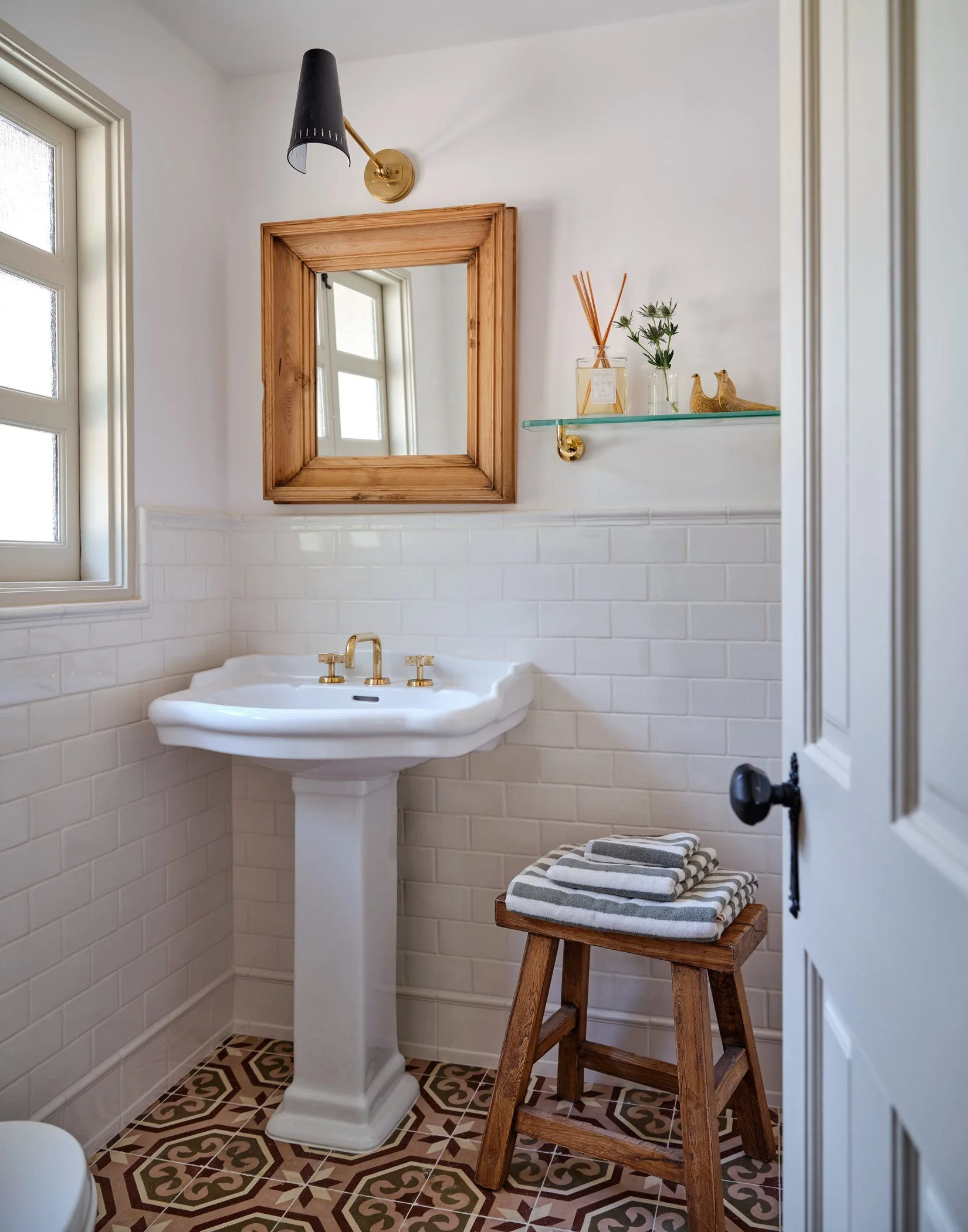 A small powder room features a white pedestal sink with brass faucets, subway tile walls, a wood-framed mirror, and a patterned tile floor.
