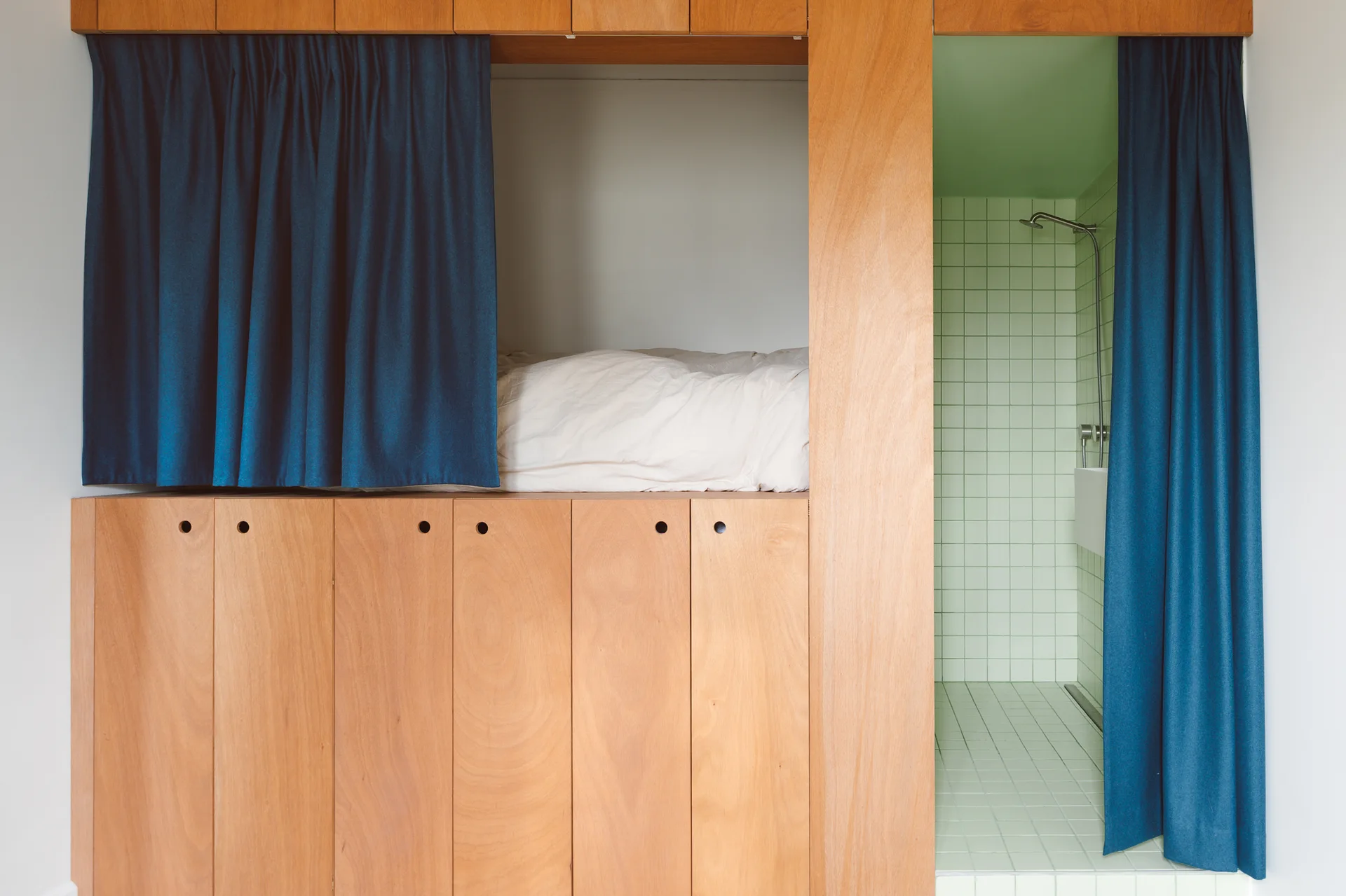 A built-in plywood unit features an elevated bed nook with blue curtains above storage cabinets, next to an integrated light green tiled shower stall.