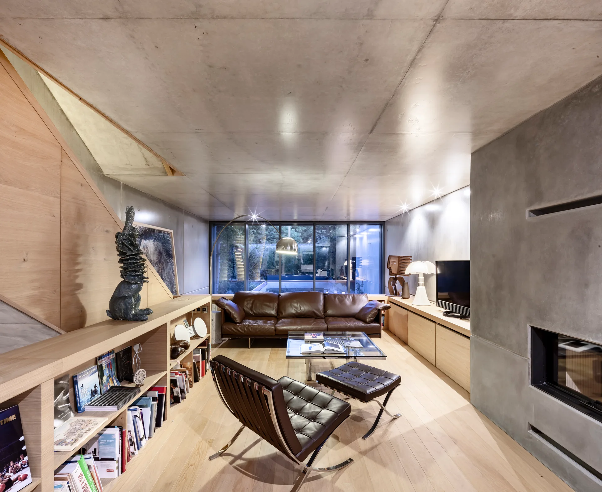 A modern living room features a brown leather sofa, a black leather lounge chair, exposed concrete walls and ceiling, and low light-wood bookshelves.