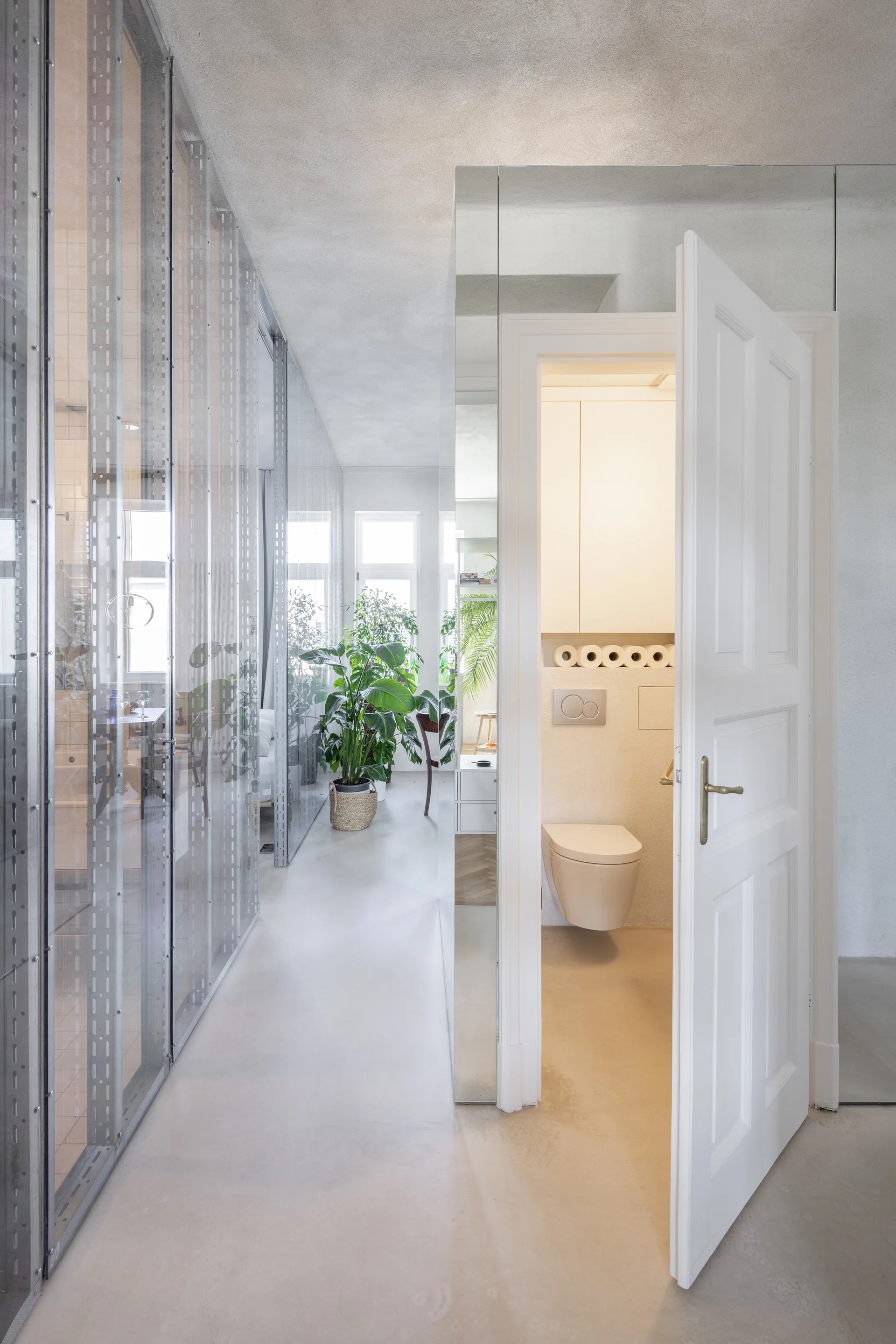 A minimalist hallway featuring a mirrored wall panel next to an open bathroom door, with an industrial glass and metal stud partition on the left.