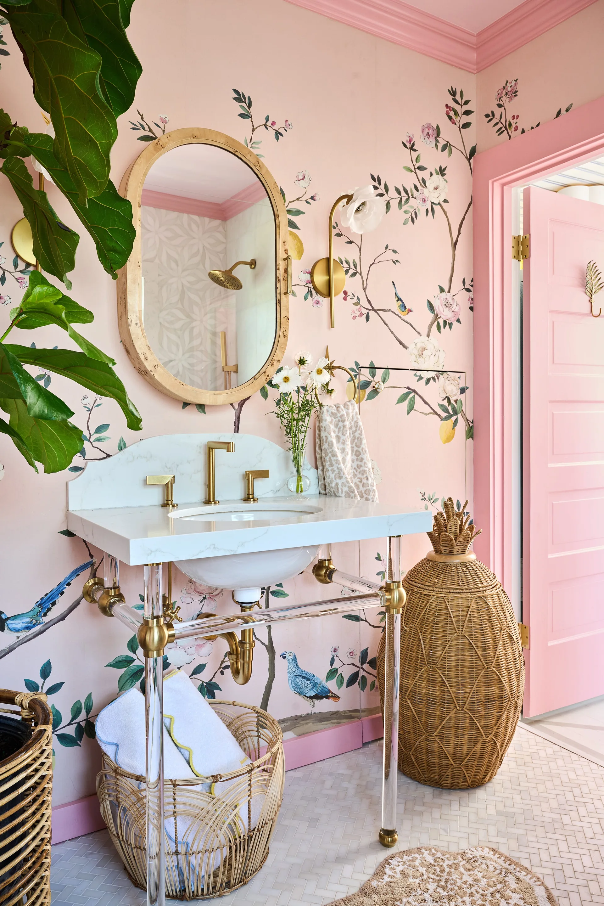 A pink bathroom features floral wallpaper, an oval wood-framed mirror, and a marble vanity with clear acrylic legs.