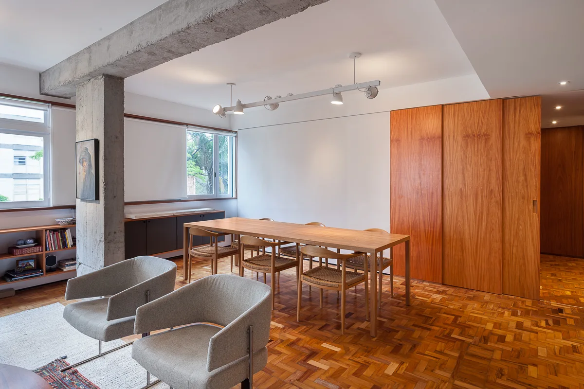 A mid-century modern dining room features a long wooden table, woven chairs, parquet flooring, a raw concrete pillar, and sliding wood wall panels.