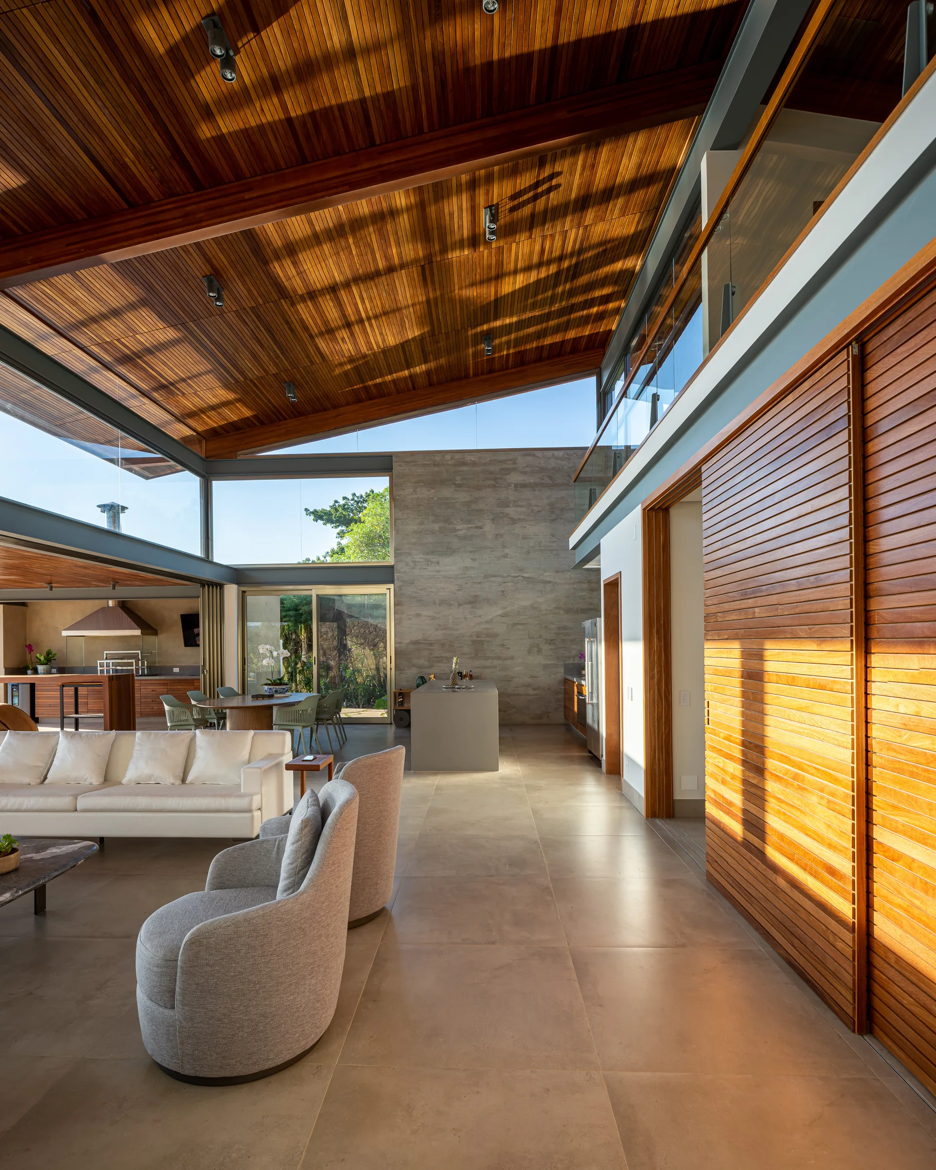 An open-concept living room features a sloped wooden ceiling, a white sofa with gray armchairs, concrete walls, and large floor-to-ceiling windows.