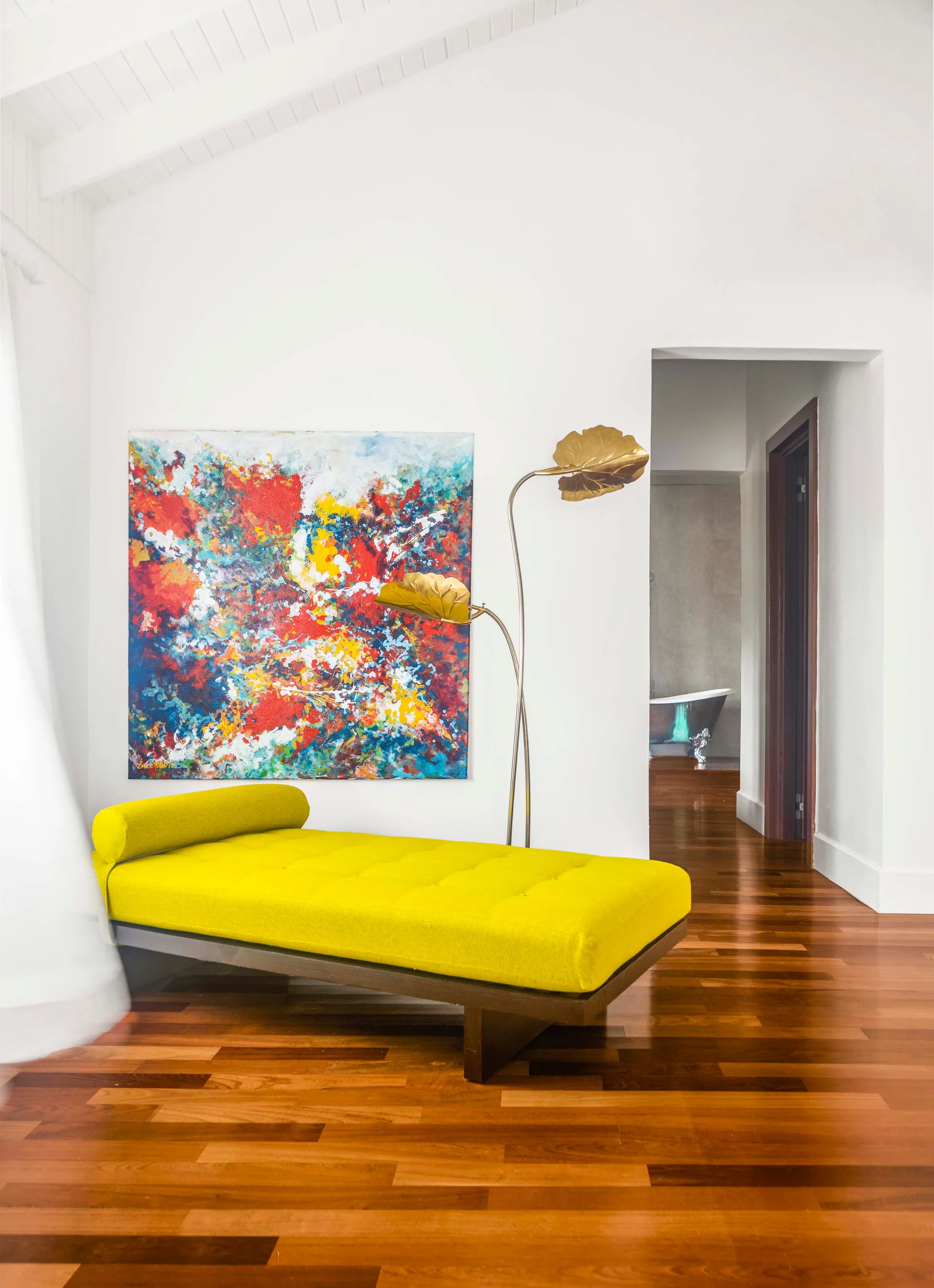 A vibrant yellow upholstered daybed sits beneath a colorful abstract canvas painting on a white wall next to a brass leaf floor lamp.