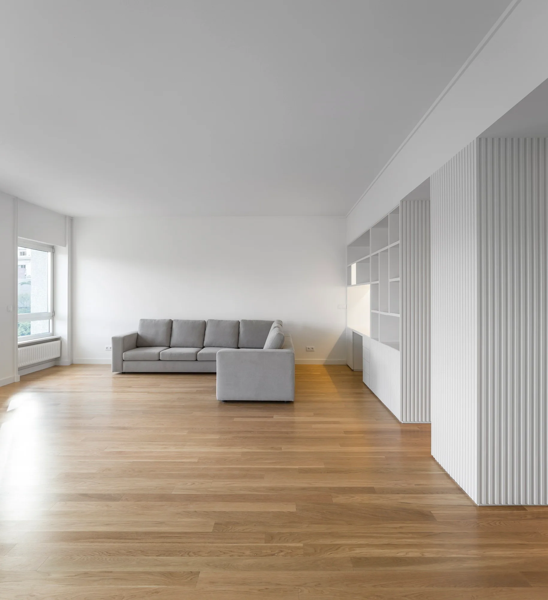 A minimalist living room featuring a grey sectional sofa, wood flooring, and white built-in shelving with vertical fluted paneling.