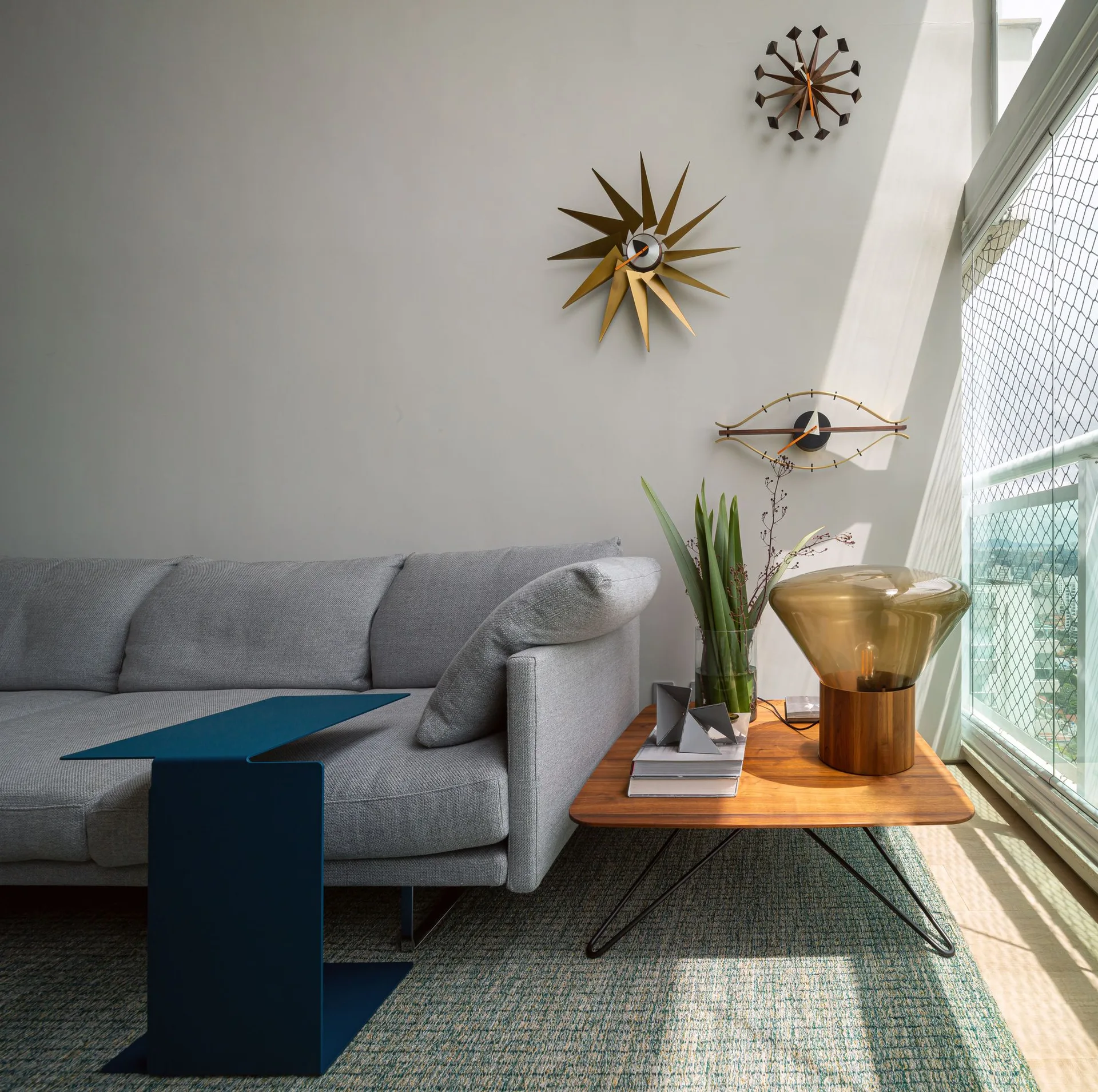 A grey sofa sits next to a wooden side table with an amber glass lamp, a blue C-table, and three starburst clocks on the wall.