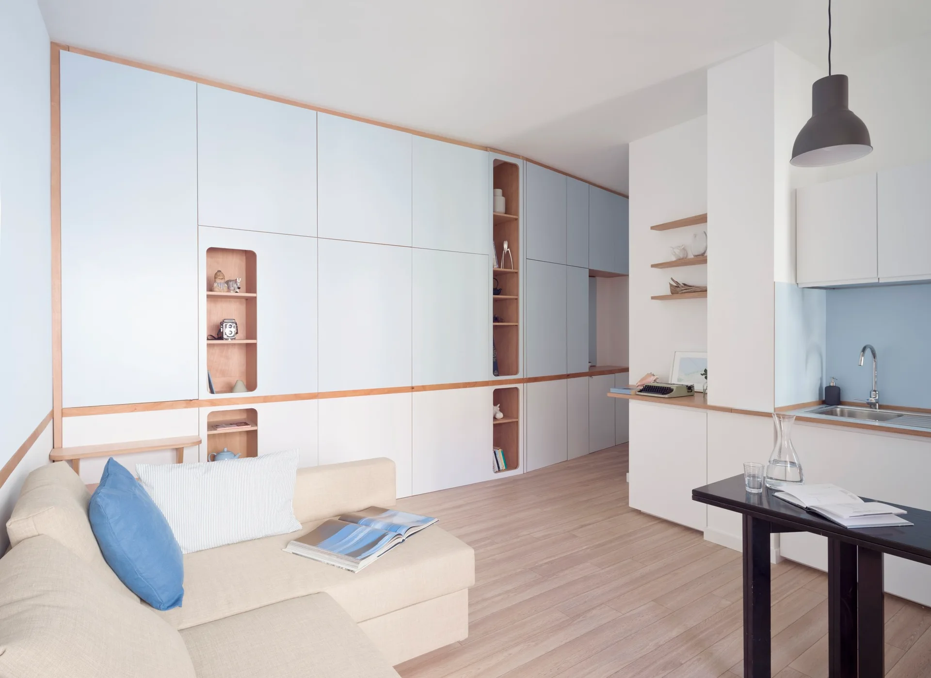 A minimalist studio apartment features a pastel blue and white custom storage wall, a cream sectional sofa, and a compact kitchen counter.