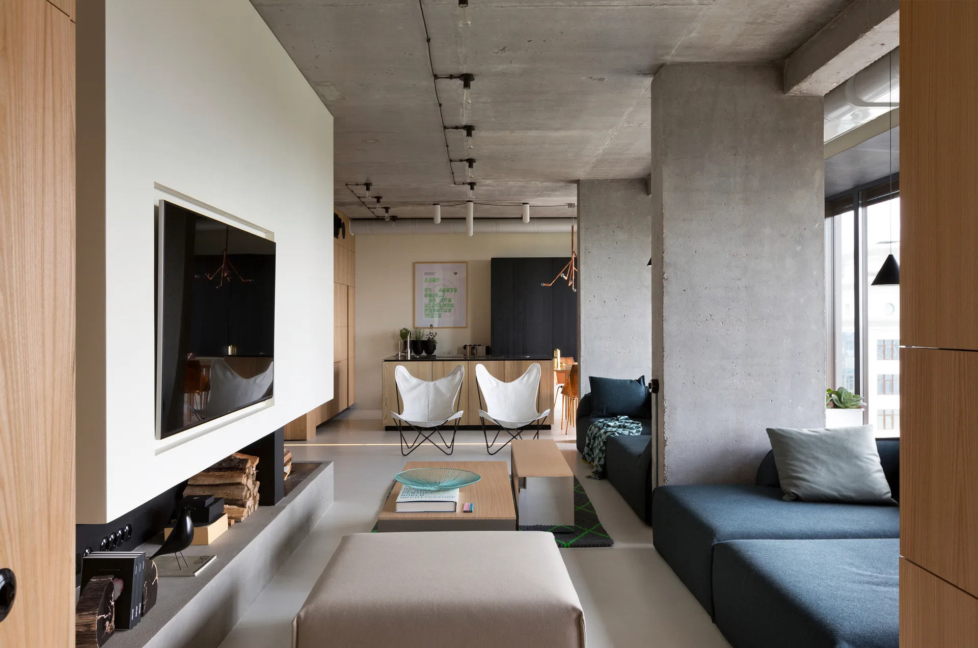 A modern open-plan living room featuring an exposed concrete ceiling, a dark sectional sofa, wood coffee tables, and two white butterfly chairs.