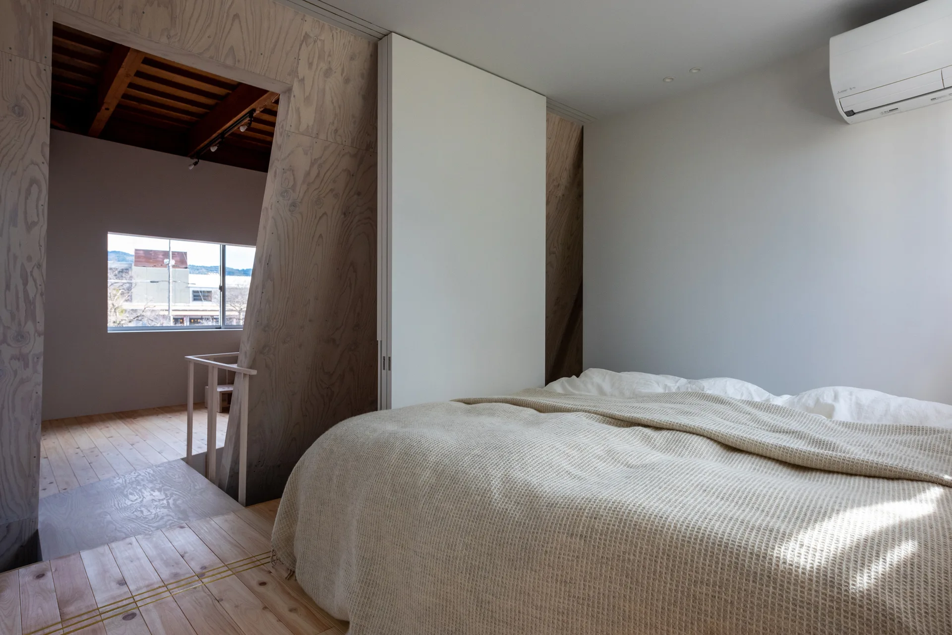 A minimalist bedroom featuring a bed with a waffle-weave blanket, natural wood flooring, a white sliding door, and an angled plywood partition wall.