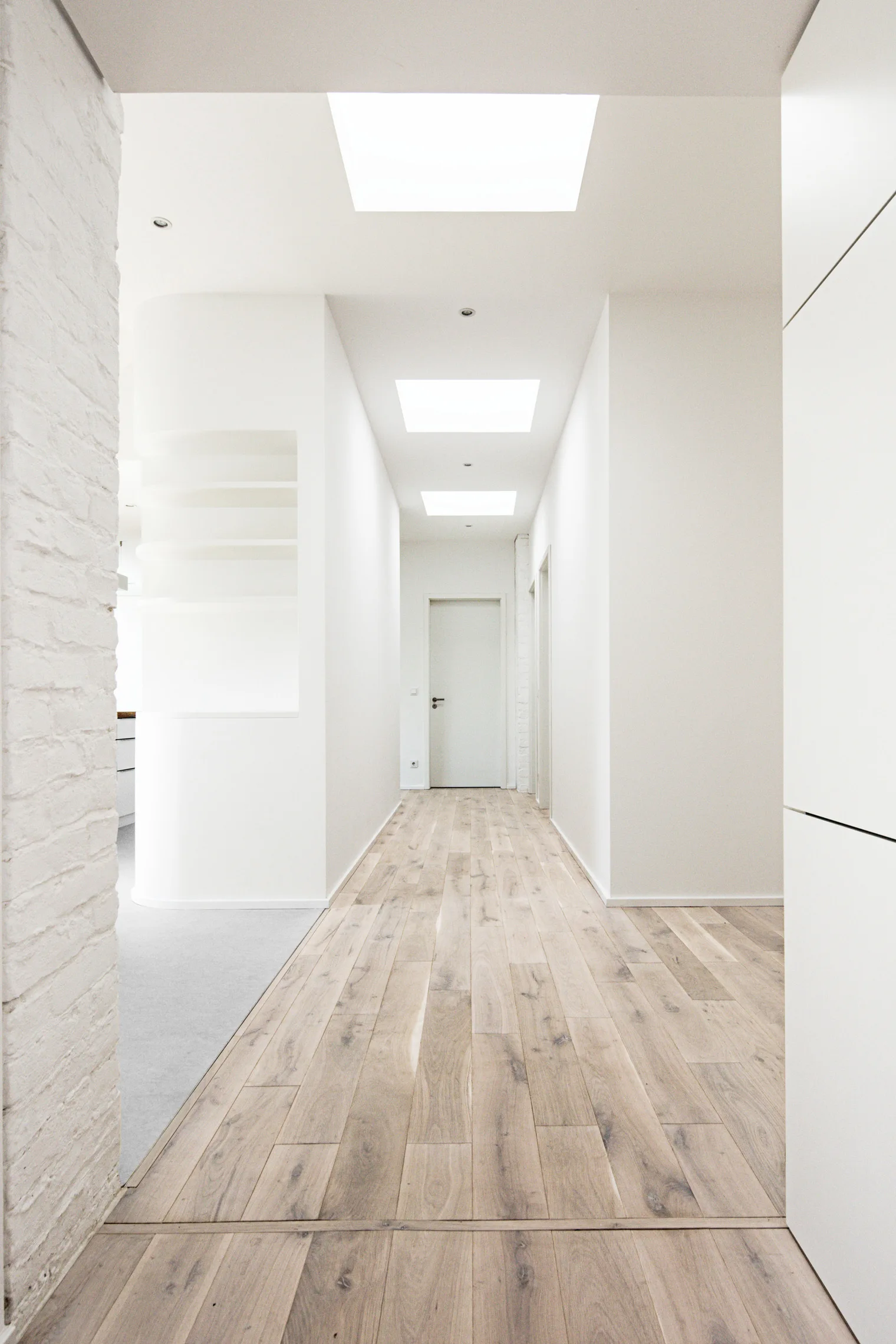 A long minimalist hallway features light wood floors, white walls with a curved niche, and square skylights illuminating the path.