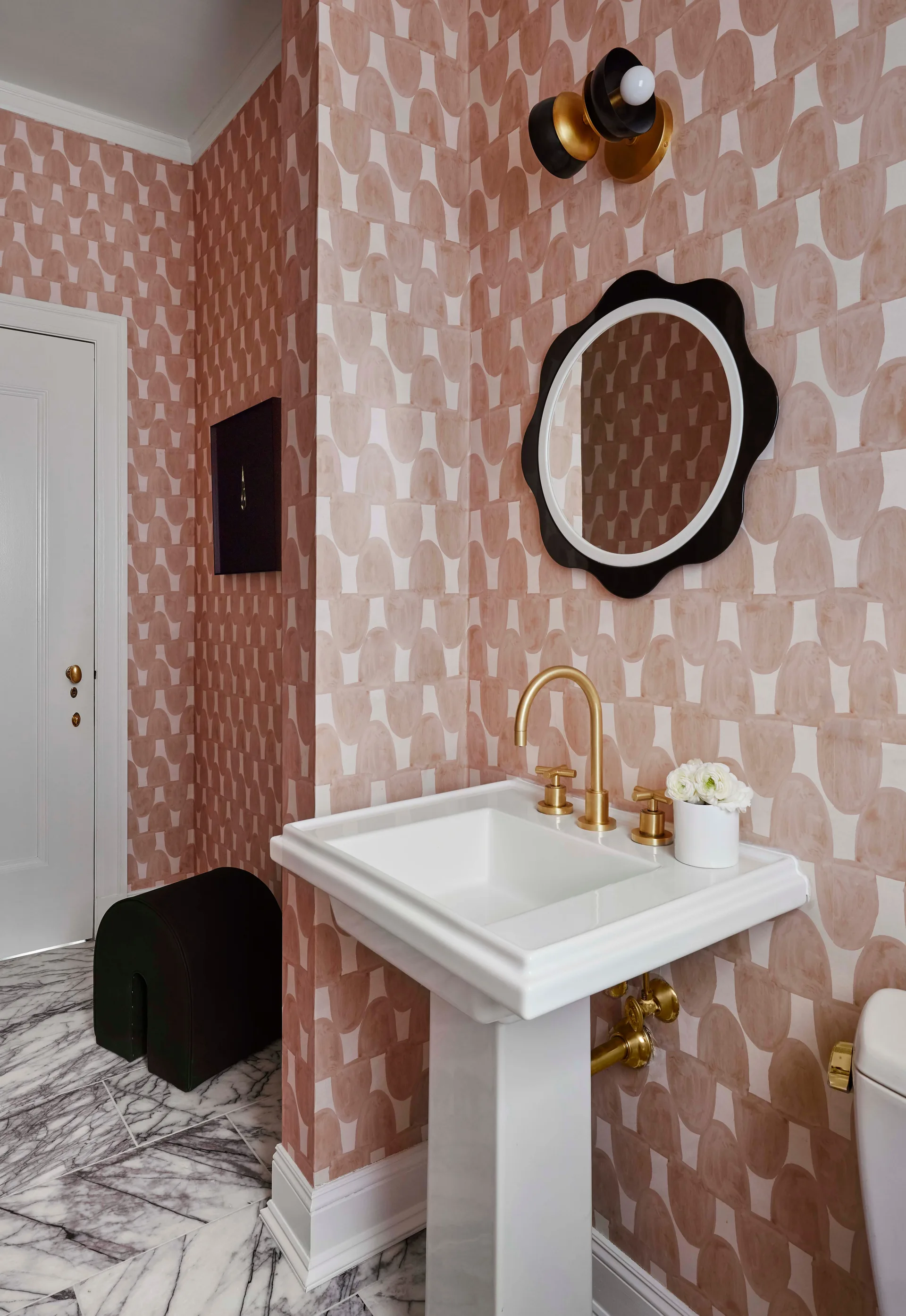 A powder room features blush pink patterned wallpaper, a white pedestal sink with brass fixtures, a black scalloped mirror, and marble flooring.