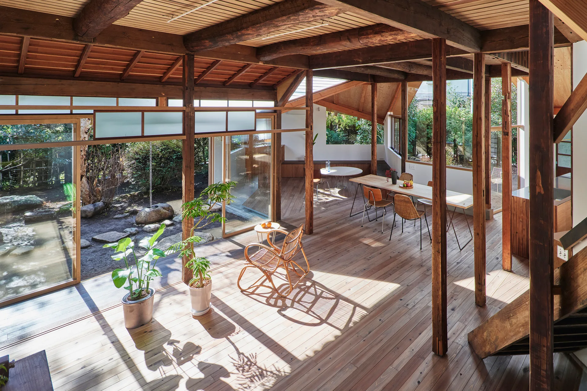An open-plan wood interior features exposed structural timber posts, a rattan rocking chair, and glass sliding doors opening to a garden.