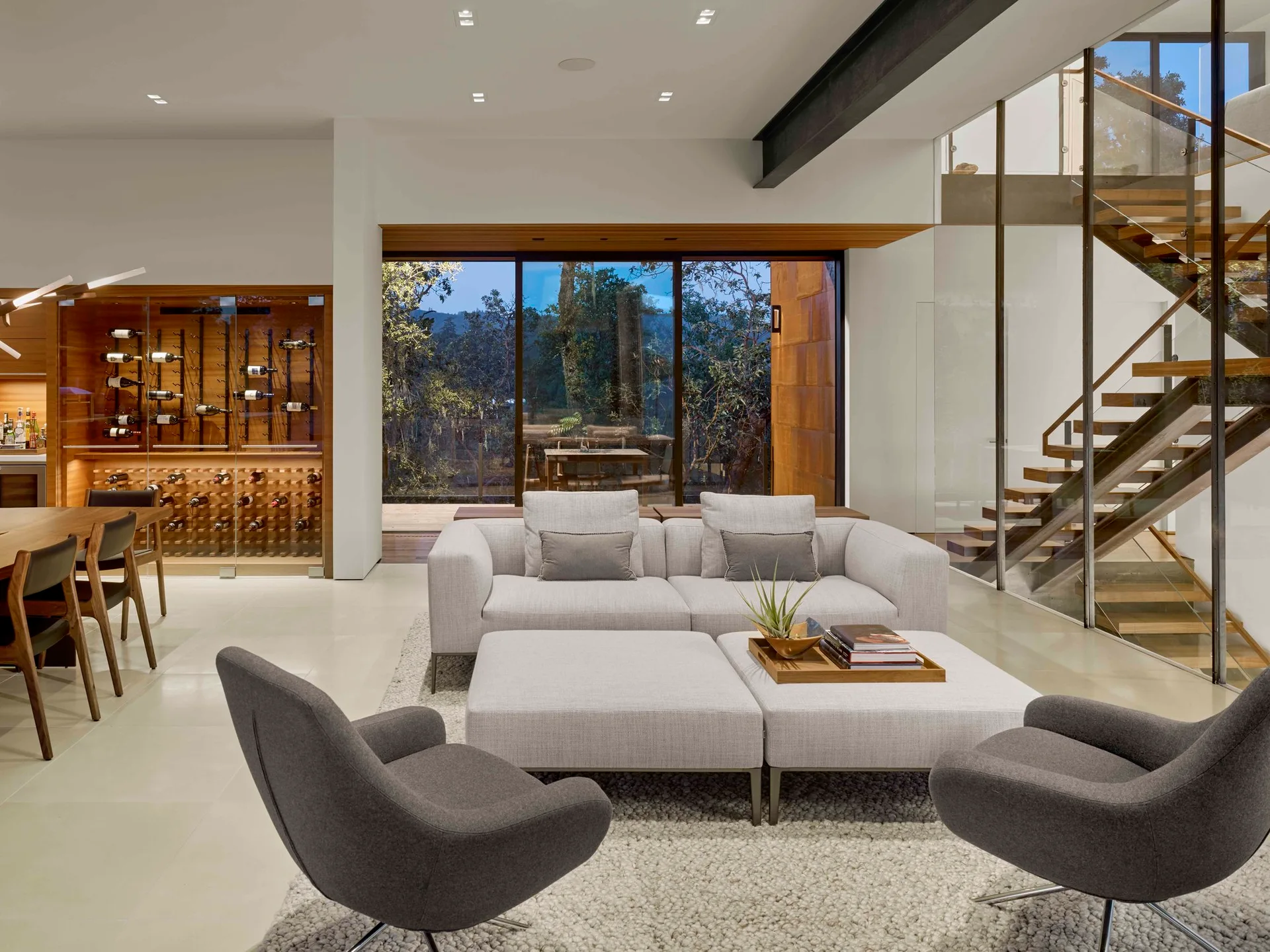 A modern open-concept living room featuring a light grey sofa, matching ottomans, a floating wooden staircase, and a glass-enclosed wine wall.
