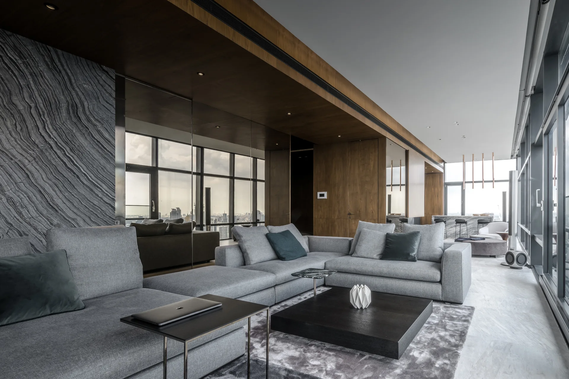A modern living room features a grey sectional sofa, a low black coffee table, wood wall paneling, and floor-to-ceiling windows.