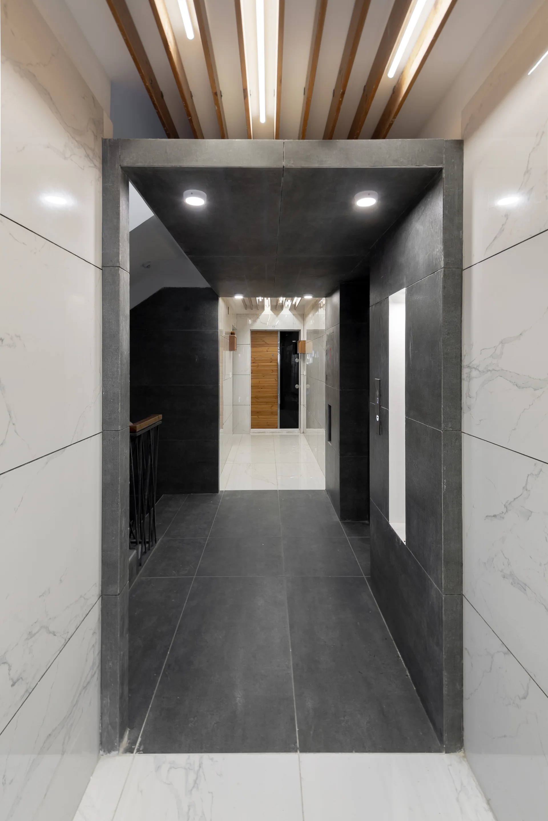 A modern hallway featuring glossy white marble wall panels, dark grey matte floor tiles, a deep grey stone archway, and a wooden slat ceiling with lights.