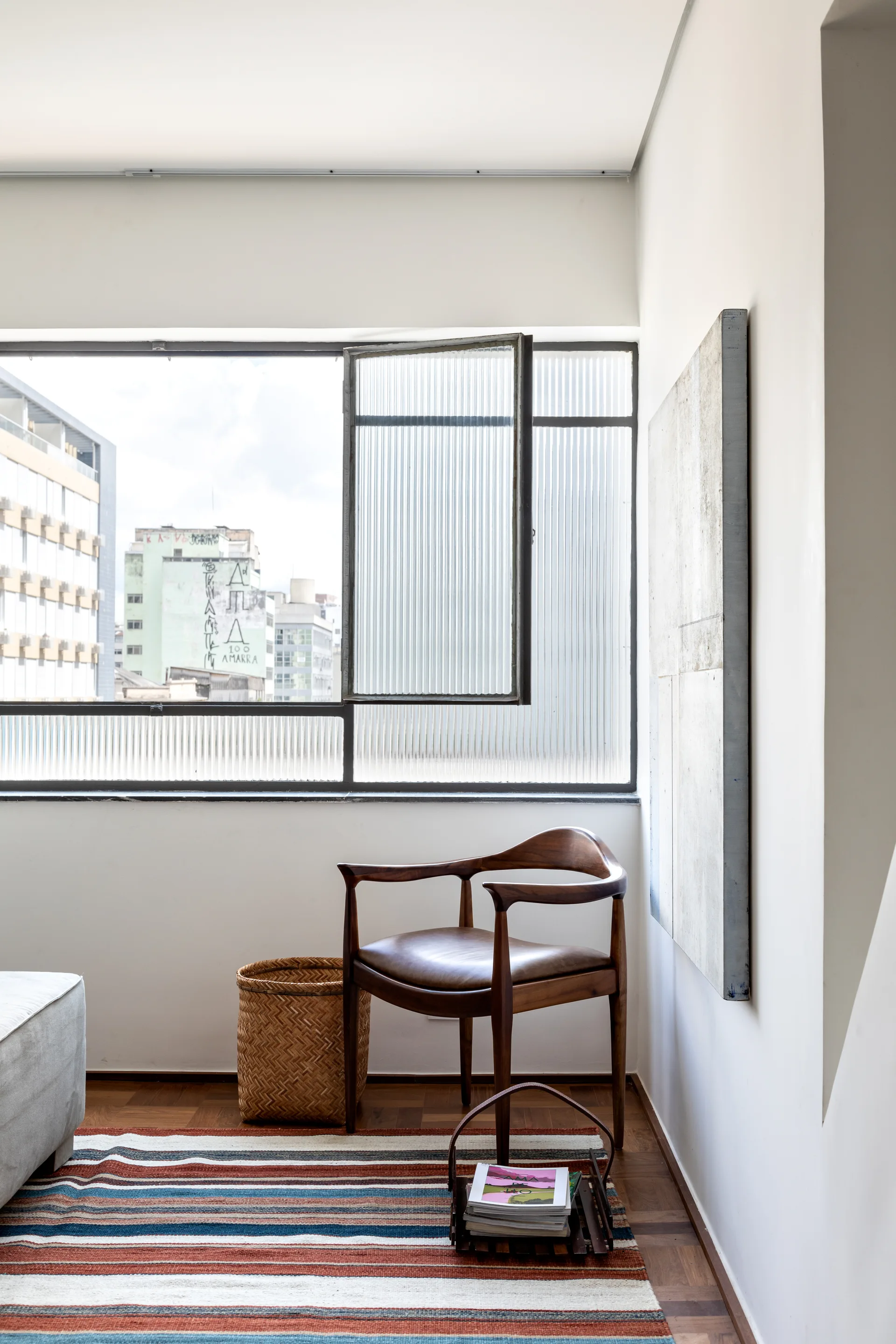 A mid-century wooden armchair sits in an apartment corner beside a window with fluted glass, a woven basket, and a striped rug.