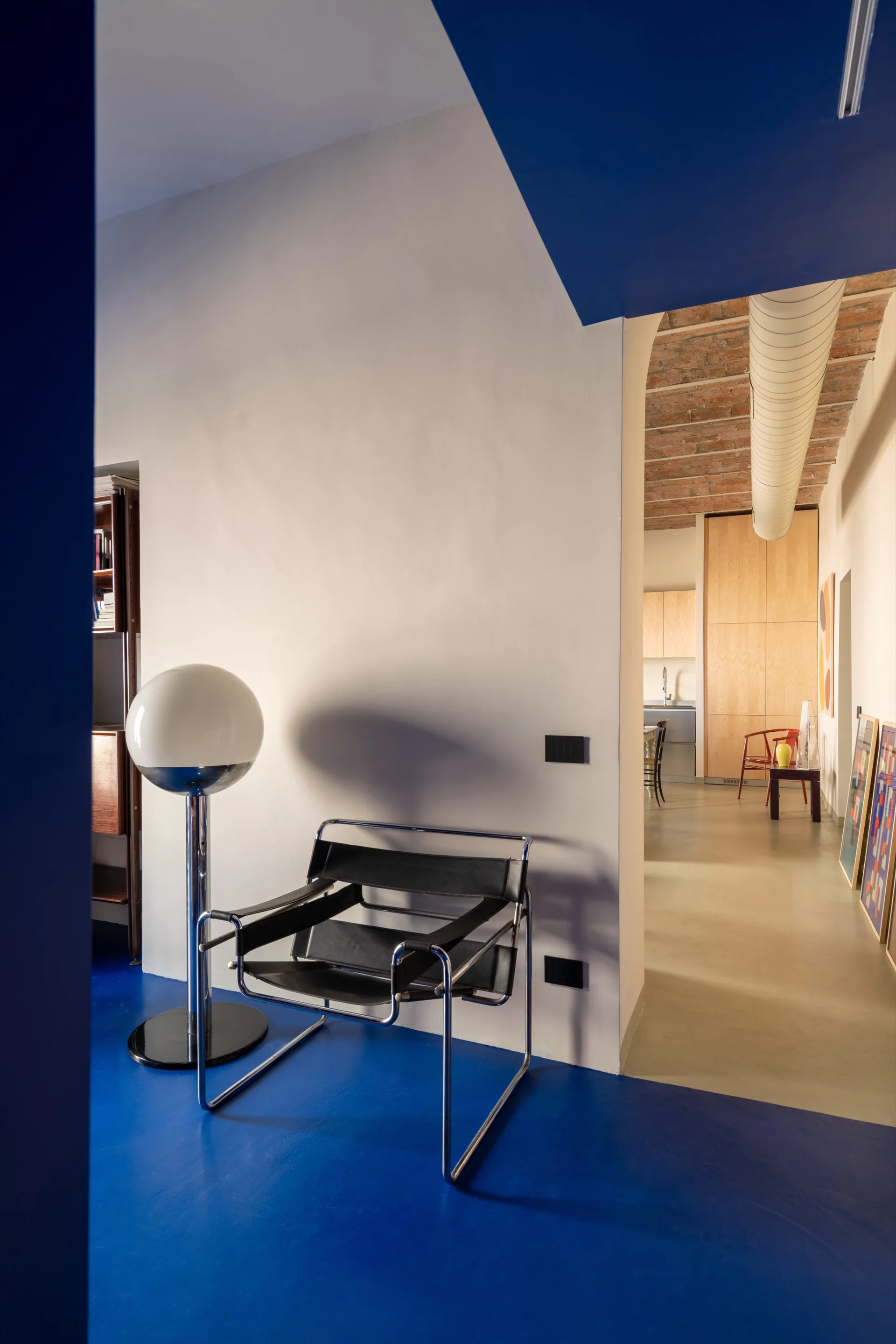 A modern living space features a vibrant blue floor, a chrome frame lounge chair with black leather straps, and an arched doorway leading to a kitchen.