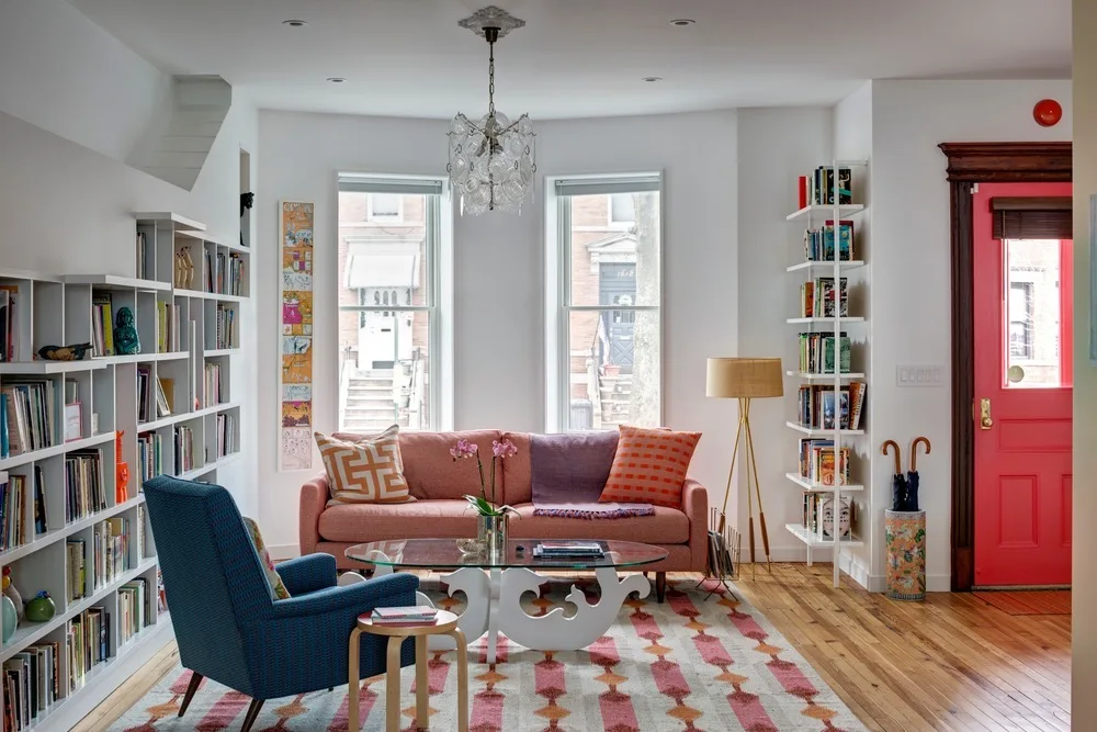 An eclectic living room features a coral sofa, a patterned blue armchair, white built-in bookshelves, and a bright pink entry door.