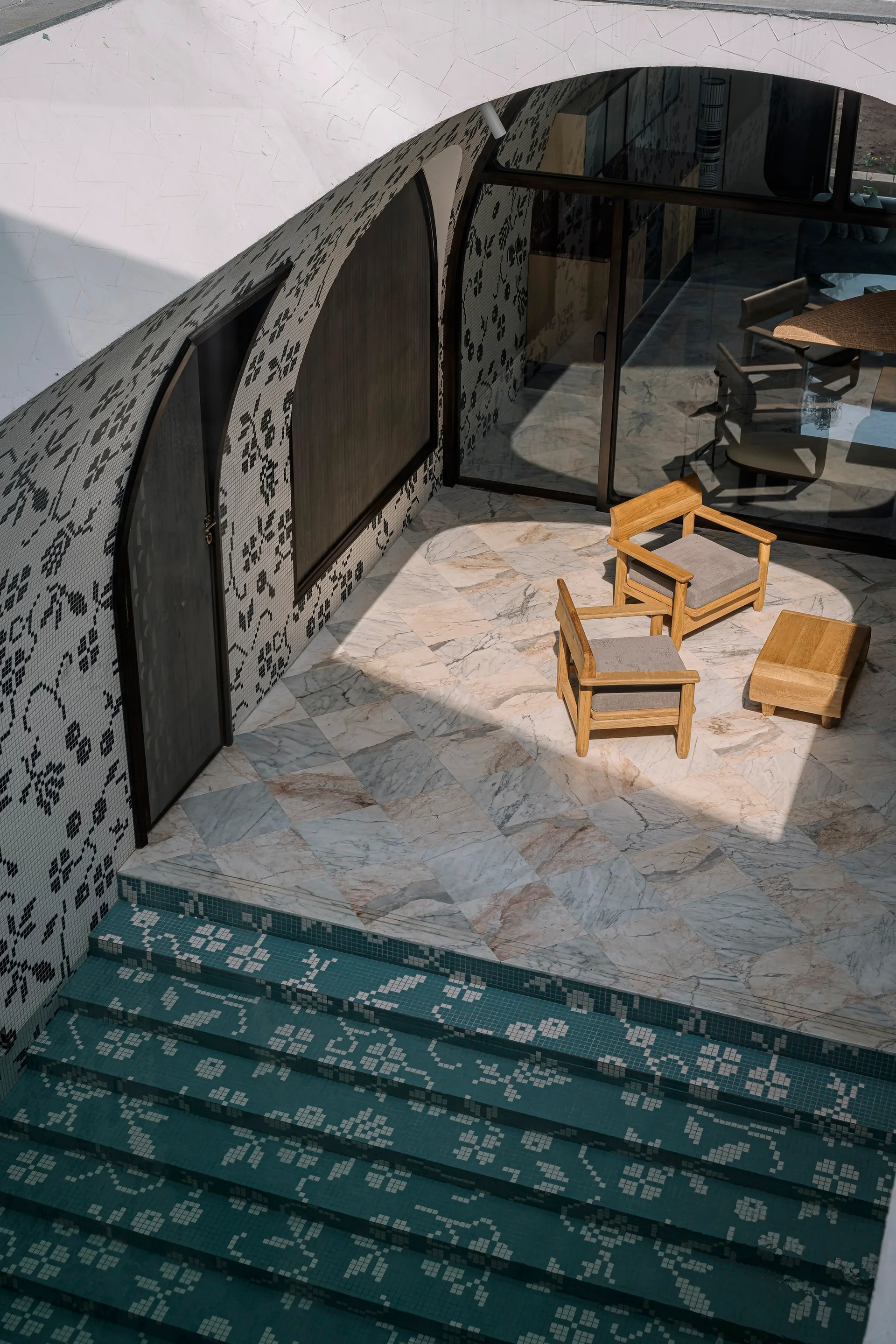 An outdoor patio features two wooden armchairs on a marble tile floor next to a teal mosaic tiled staircase and arched walls.