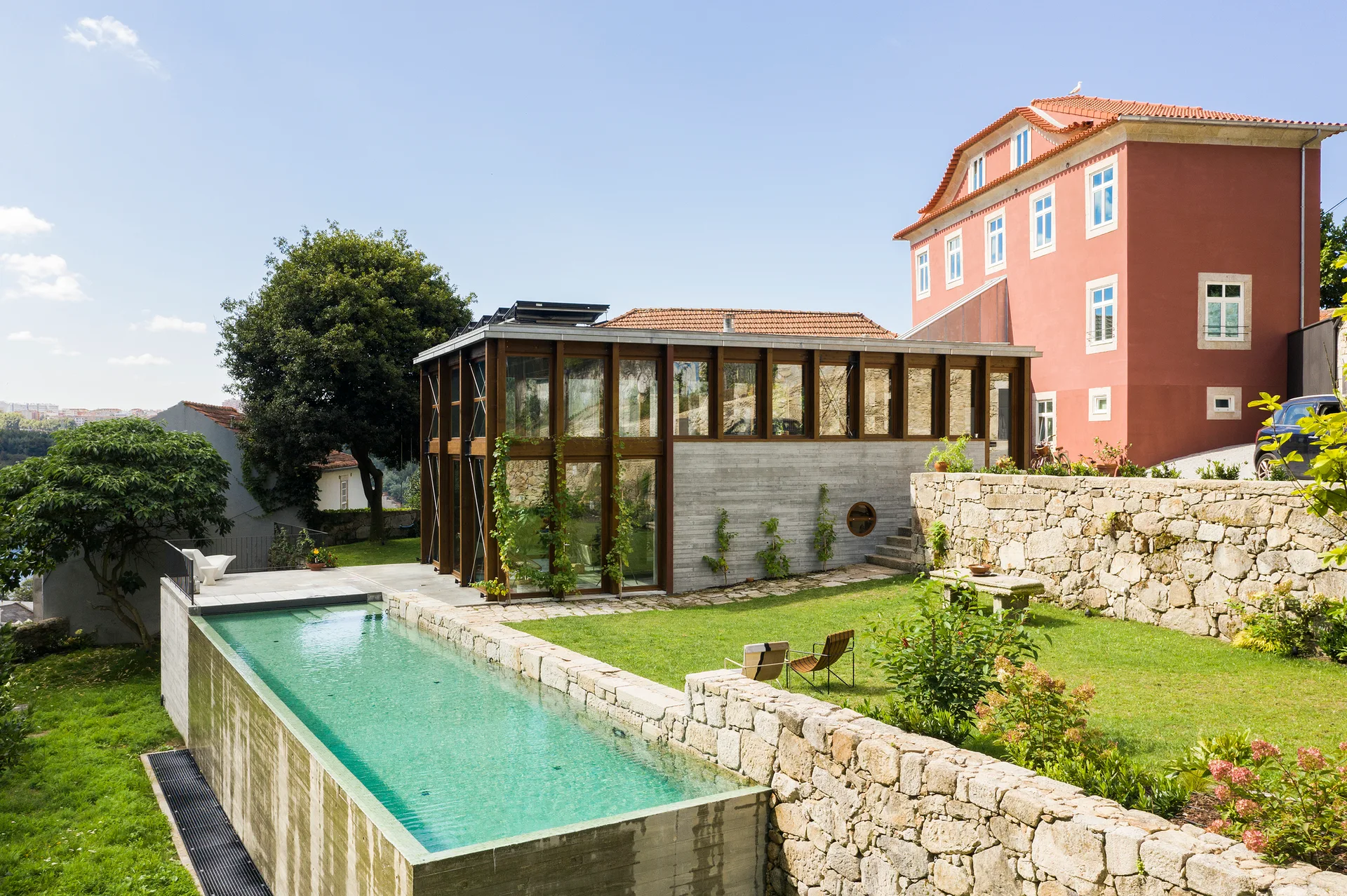 A rectangular swimming pool with a concrete side sits next to a green lawn with a modern glass pavilion and stone retaining walls under a blue sky.