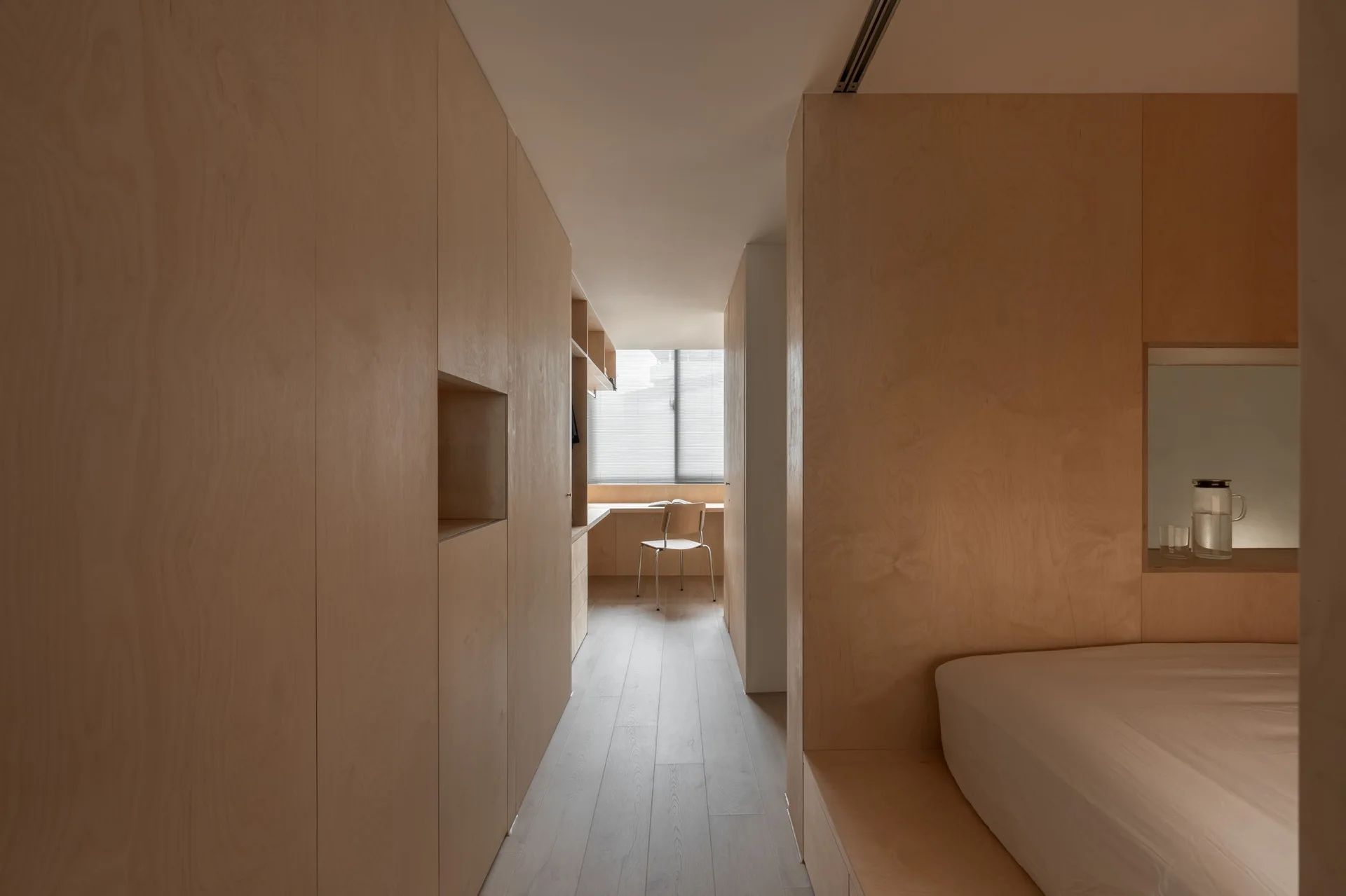 A minimalist bedroom interior lined with light wood veneer cabinetry features a built-in desk at the far end and an integrated bed platform.