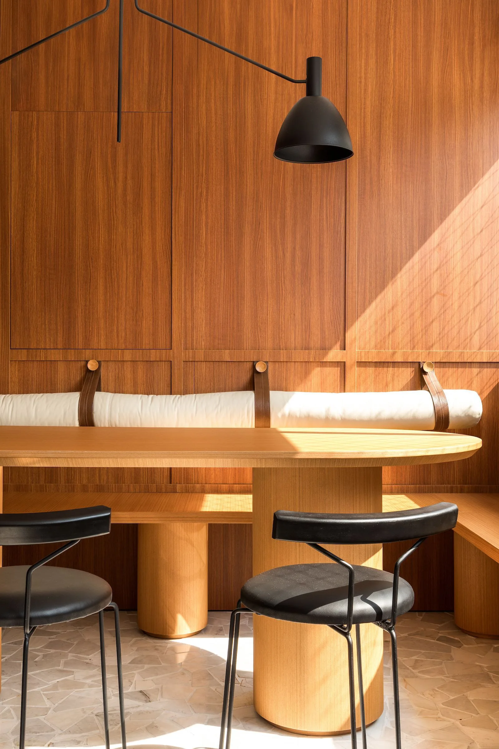 A modern dining nook features wood paneled walls, a light wood table, and a built-in bench with an off-white bolster cushion held by leather straps.