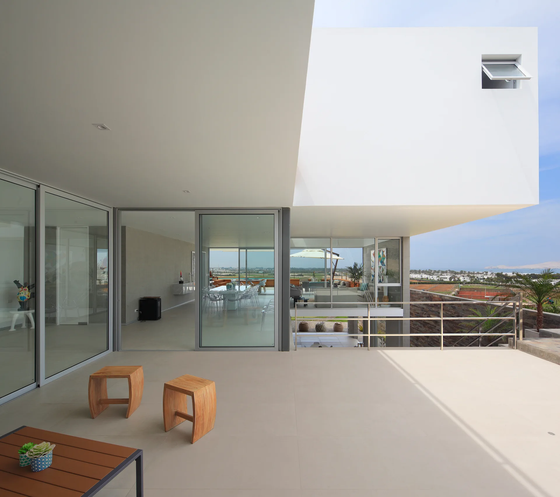 A minimalist modern patio features large glass sliding doors, curved wooden stools, and a dramatic white cantilevered ceiling overhang.
