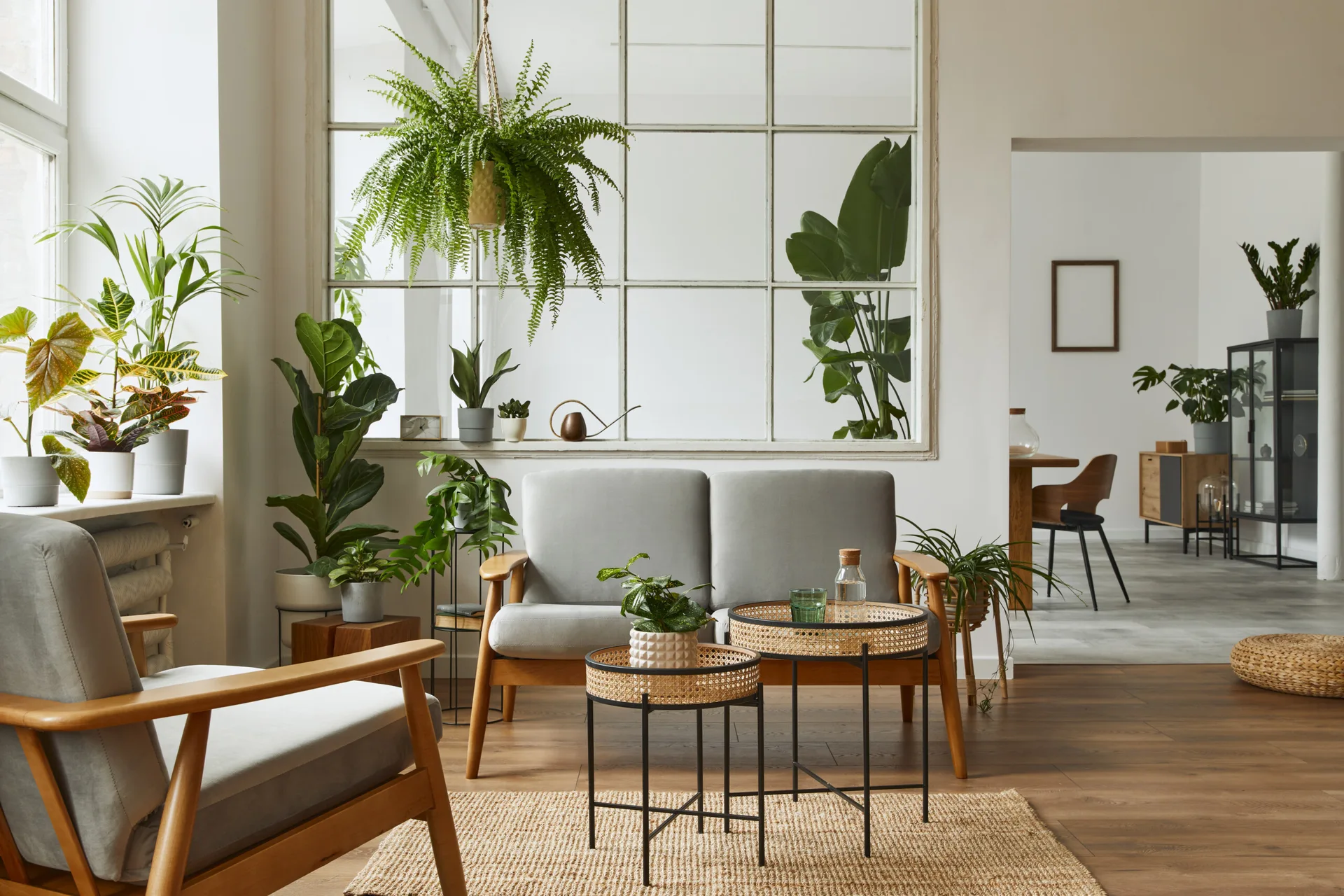 A bright living room features a grey sofa and armchair with wooden frames, rattan nesting coffee tables, a large grid window, and numerous potted plants.