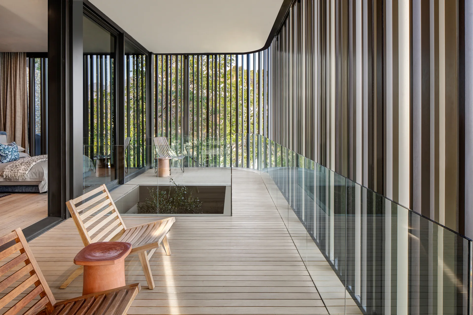 A modern outdoor terrace with a light wood deck, slatted wooden lounge chairs, a glass safety railing, and a dark vertical slat privacy screen.