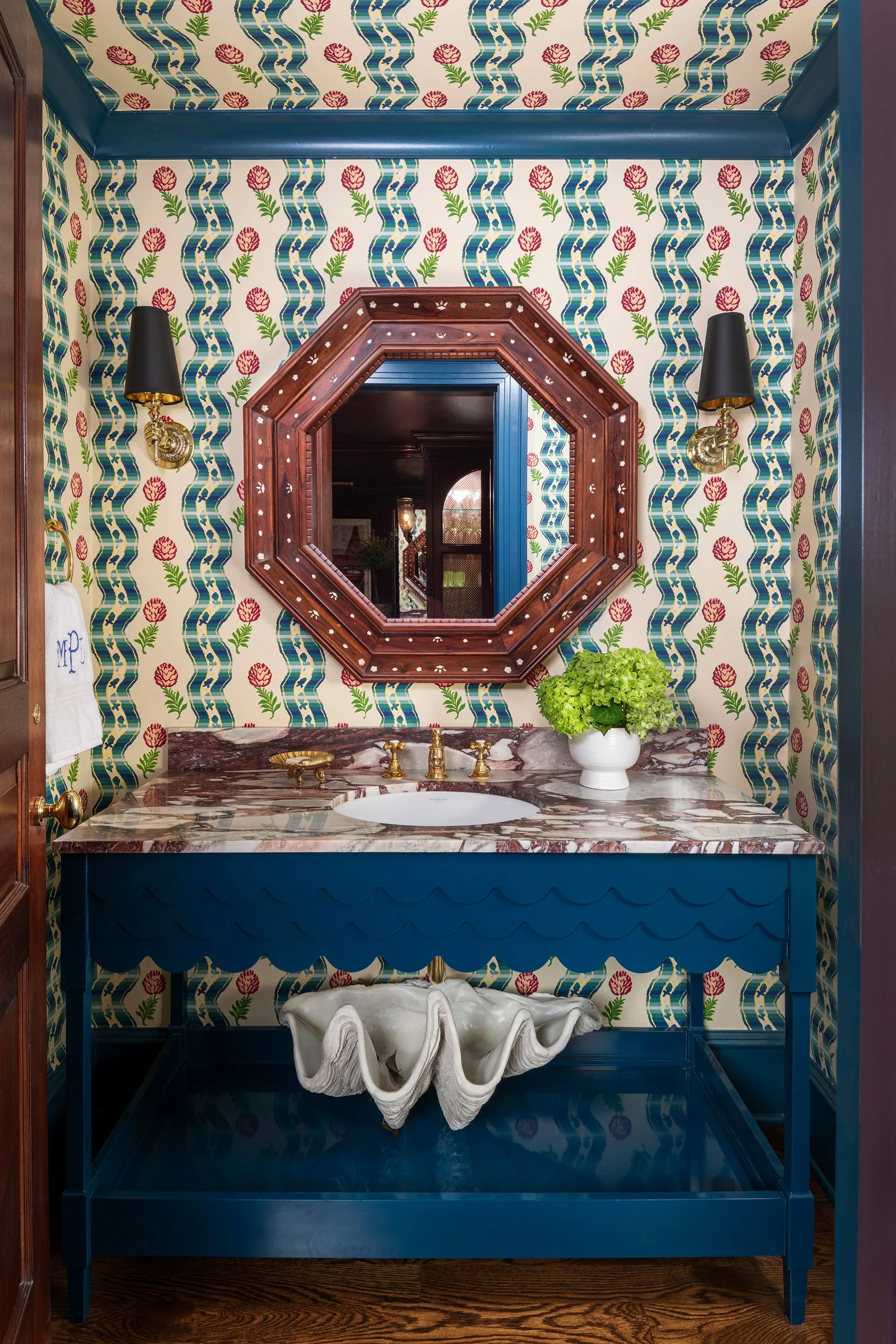 A powder room features a blue scalloped vanity with a marble countertop, an octagonal wood mirror, and floral-and-plaid patterned wallpaper.