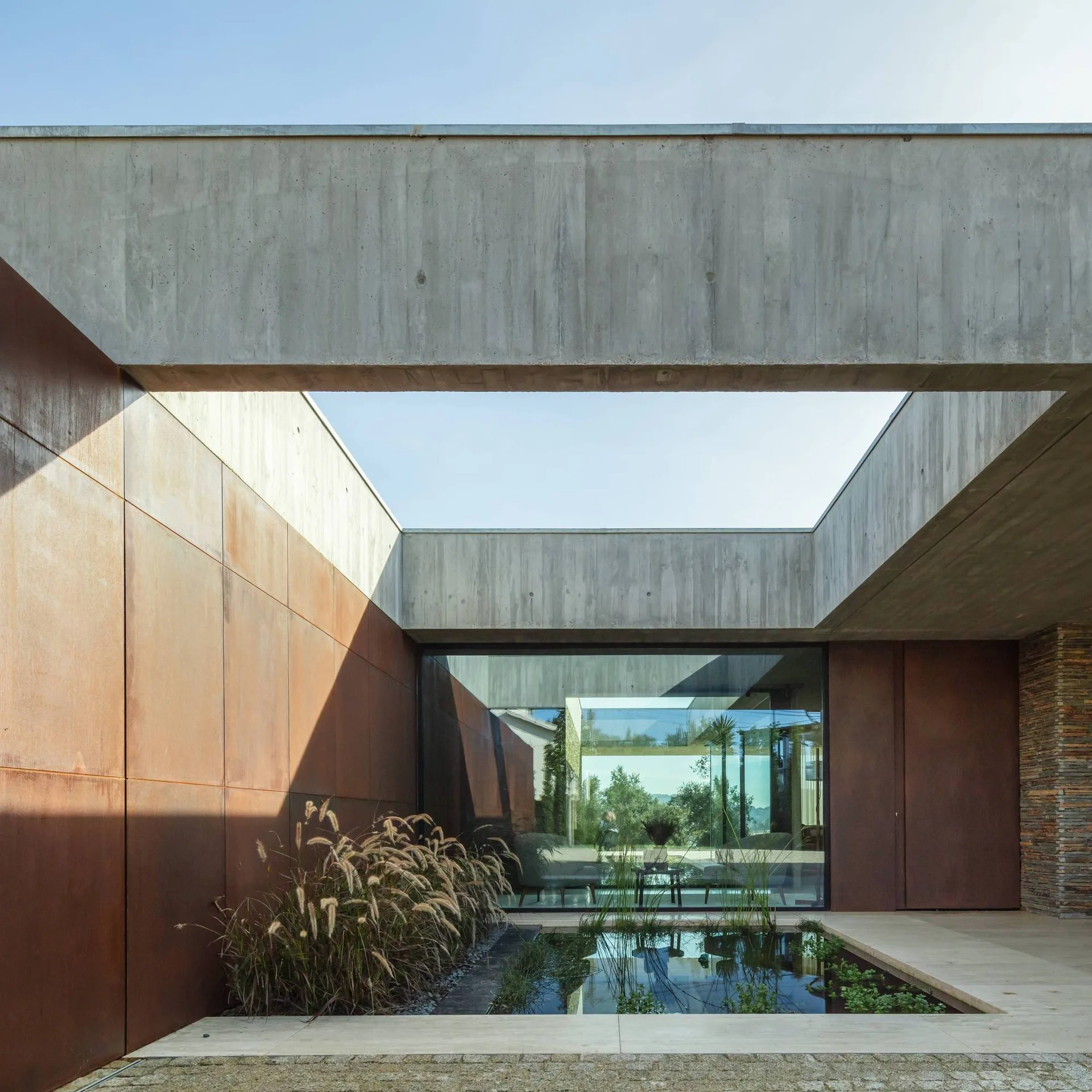 A minimalist courtyard featuring a concrete overhead frame, rusted metal wall panels, and a small pond with ornamental grasses.