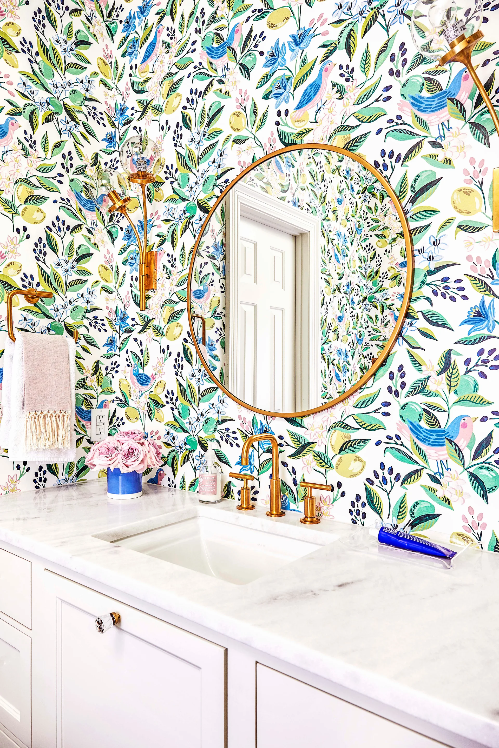 A bathroom vanity featuring a white marble countertop, a round gold-framed mirror, and colorful floral wallpaper with bird patterns.