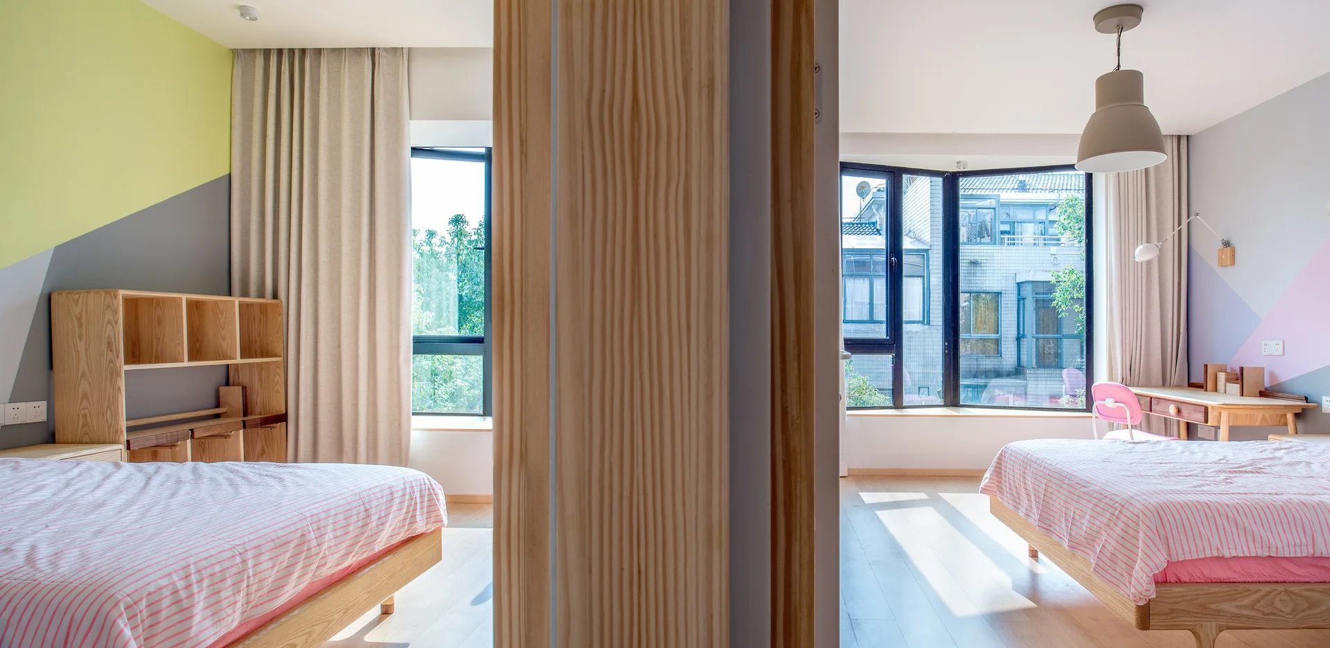 A shared kids' bedroom split by a central wooden column features twin beds with pink striped bedding, light wood furniture, and geometric painted accent walls.