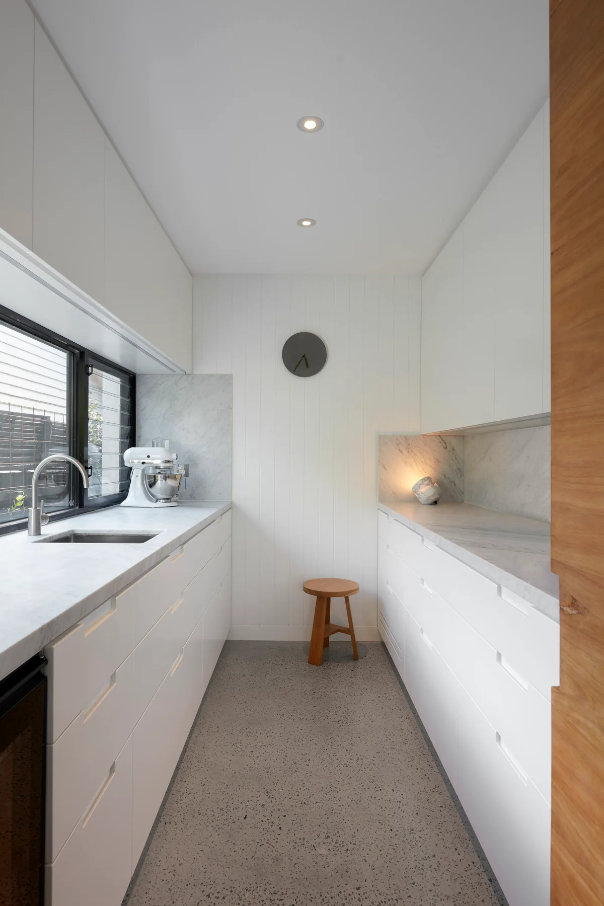 A narrow white galley kitchen features flat-panel cabinets, marble countertops, a small wooden stool, and a circular gray clock on a paneled wall.