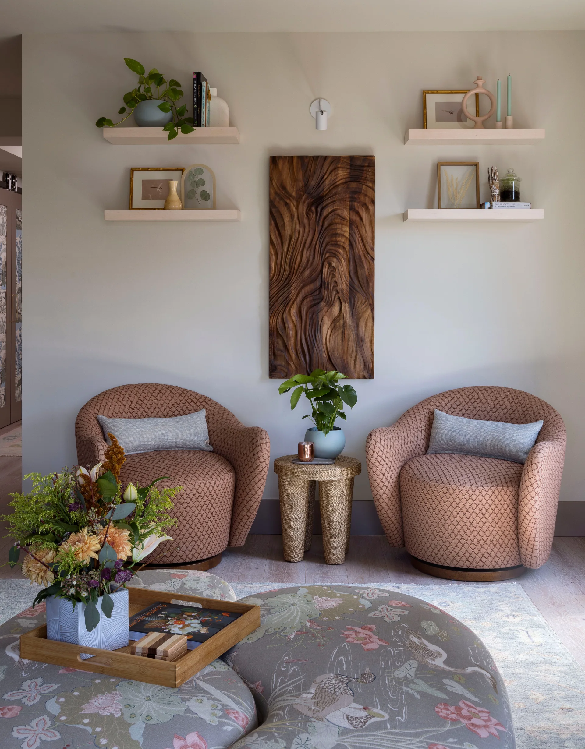 Two peach diamond-patterned armchairs flank a rope side table beneath floating shelves and a vertical carved wood sculpture.