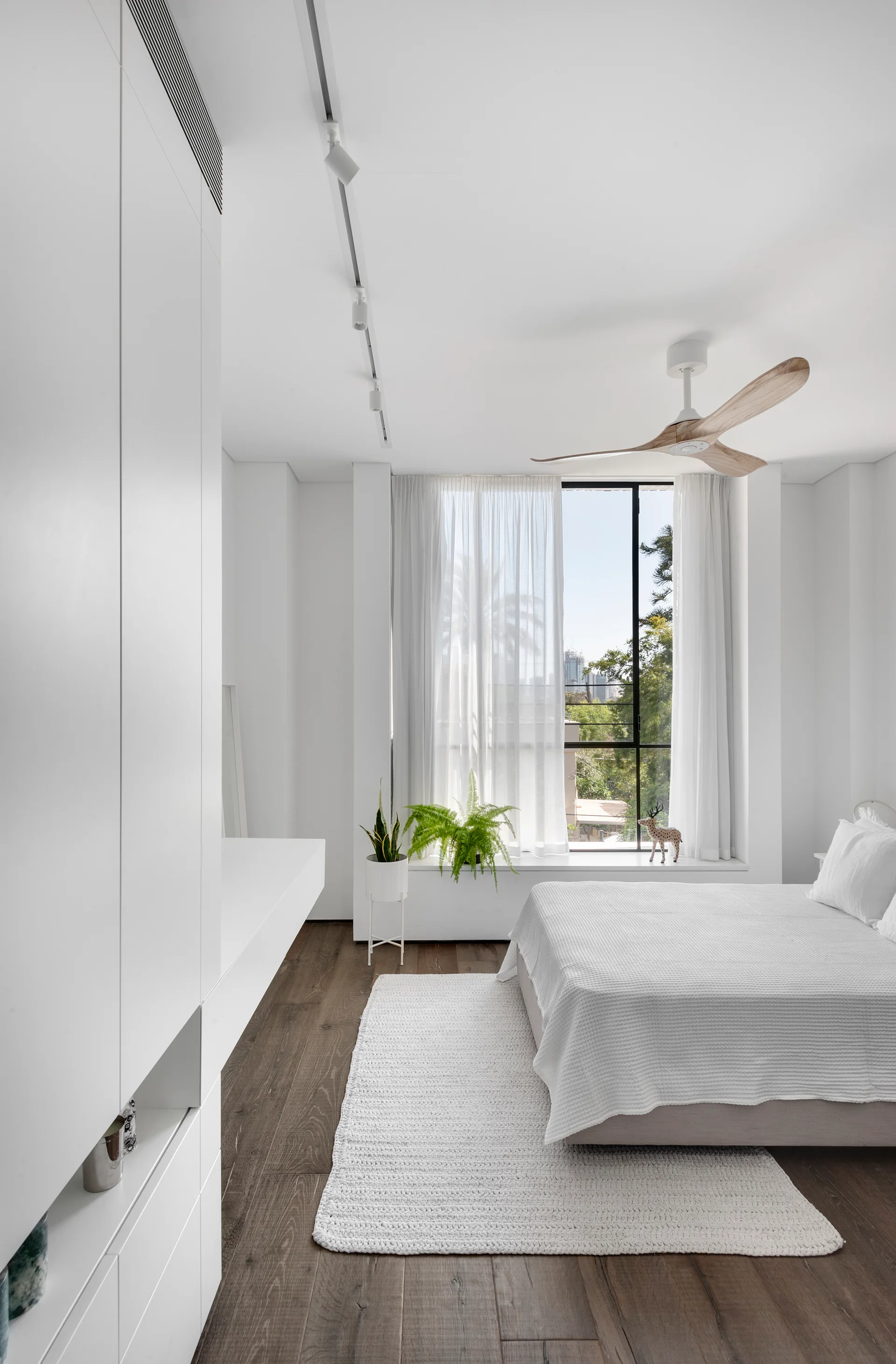 A minimalist white bedroom features floor-to-ceiling built-in cabinets, a bed with textured linens on a dark wood floor, and a wooden ceiling fan.