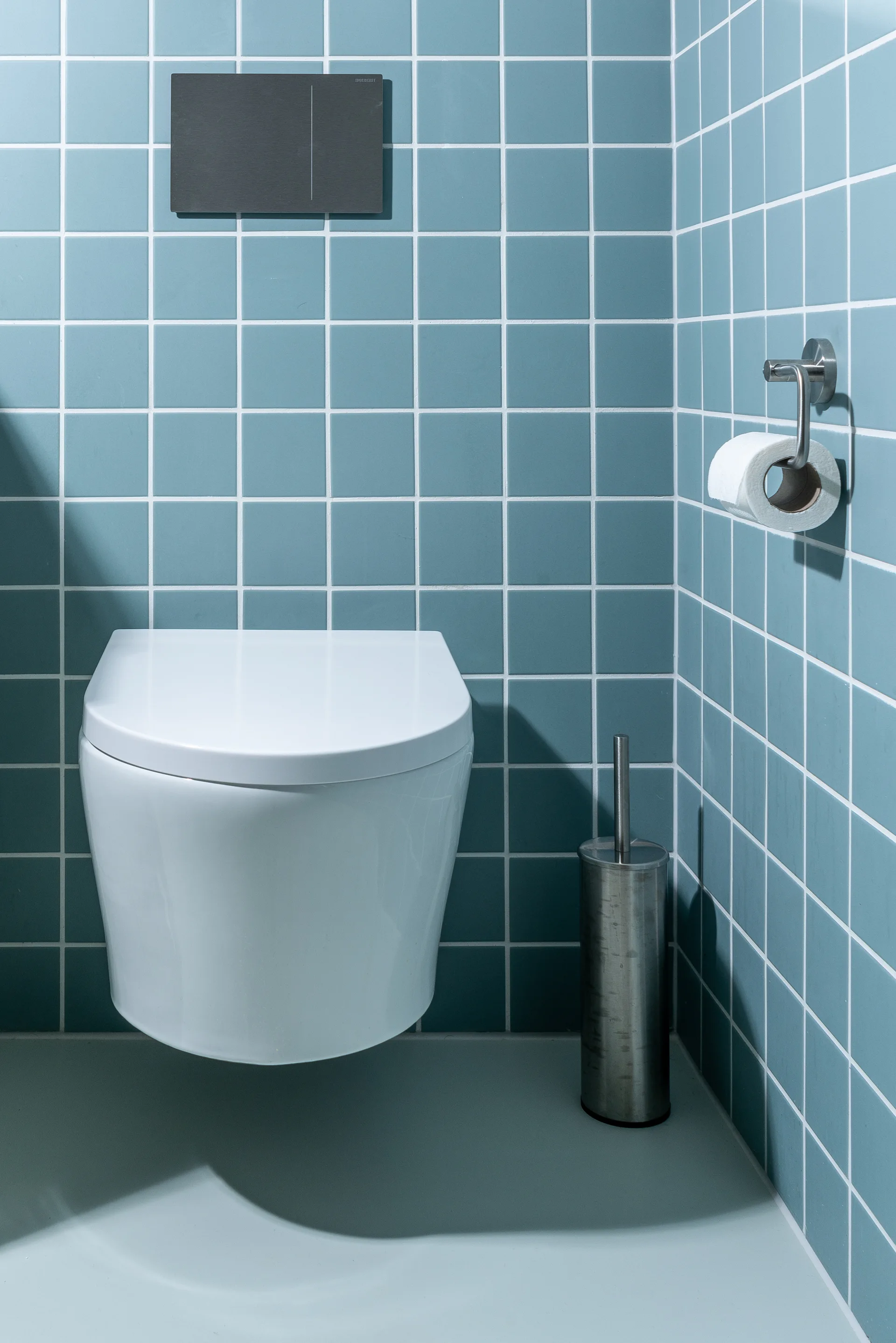 A white wall-mounted toilet sits in a bathroom corner covered in blue square grid tile with a dark grey flush plate.