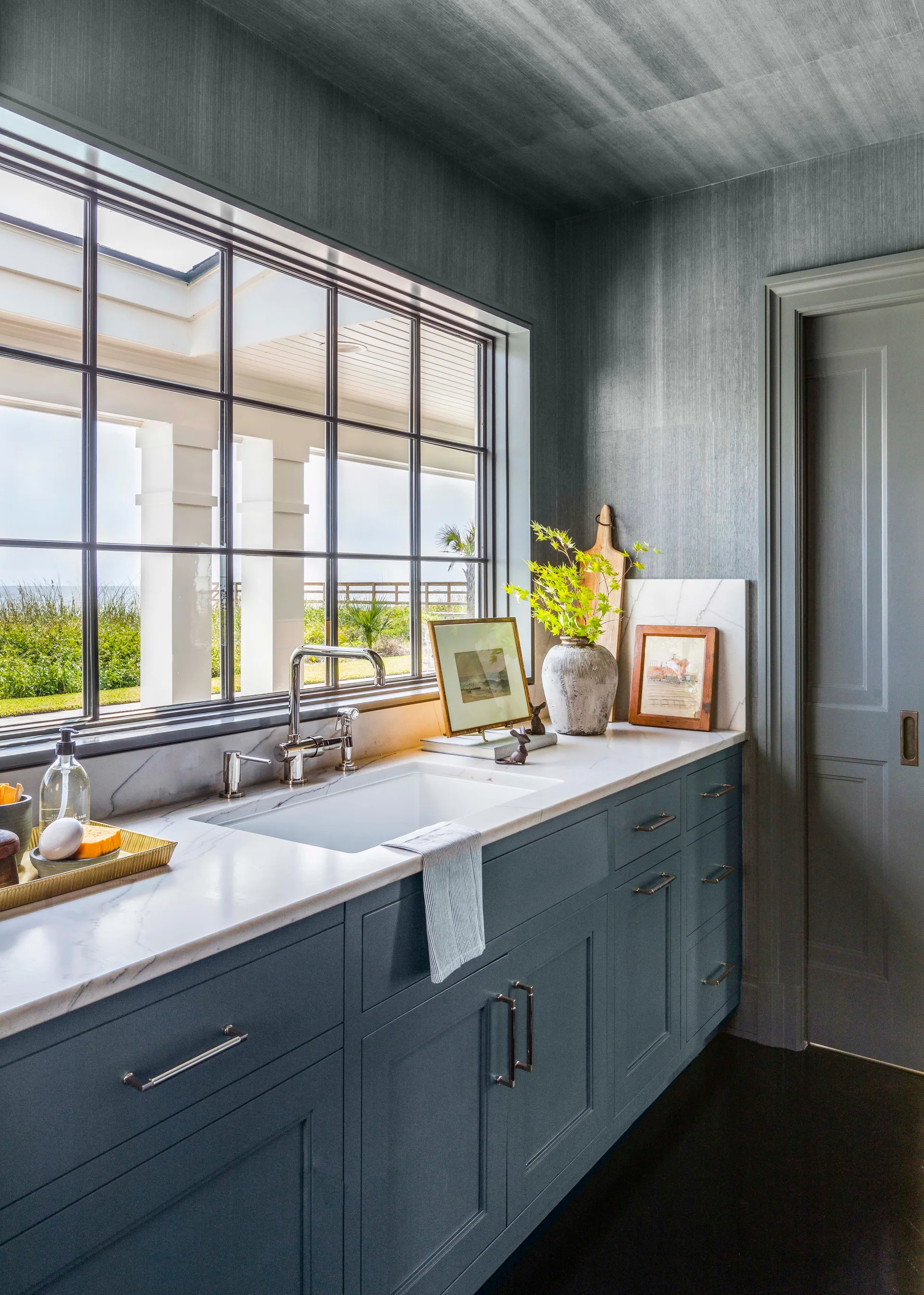 A bright kitchen area features slate blue base cabinets under a white marble countertop with a sink, set against textured gray wallpapered walls.