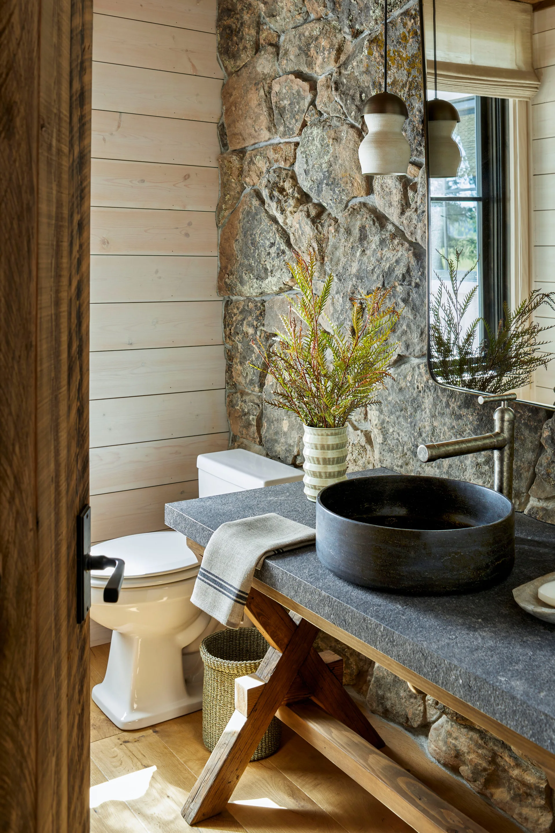 A rustic powder room featuring a stone masonry feature wall, horizontal light wood paneling, and an X-frame vanity with a dark vessel sink.