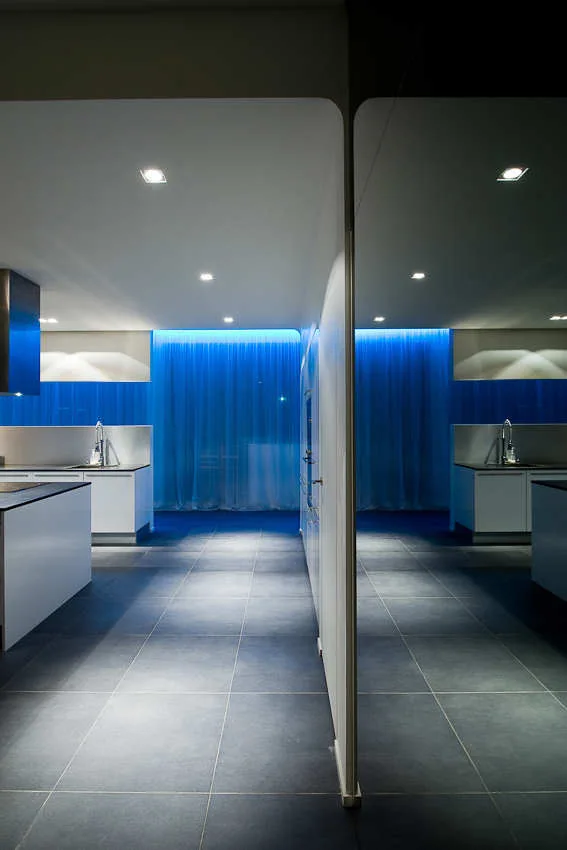 A minimalist modern kitchen with white cabinetry, dark gray tile floors, and a vibrant blue backlit curtain in the background.