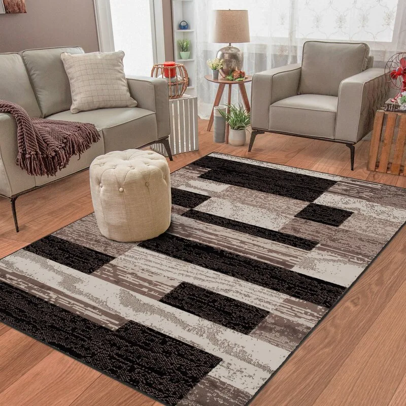 A gray sofa and armchair face a large black and brown geometric rug with a small tufted cream ottoman in a living room.