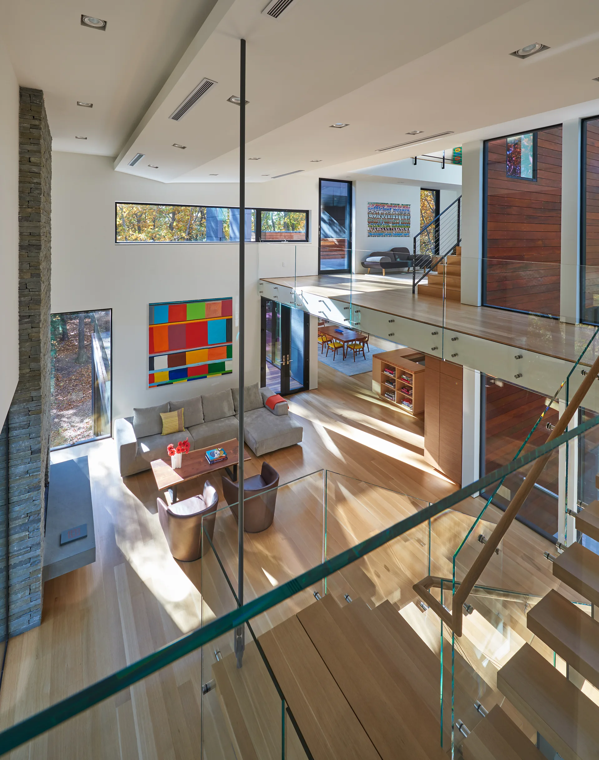 A modern multi-level living room features a grey sectional sofa, brown leather armchairs, and a wooden staircase with glass railings.