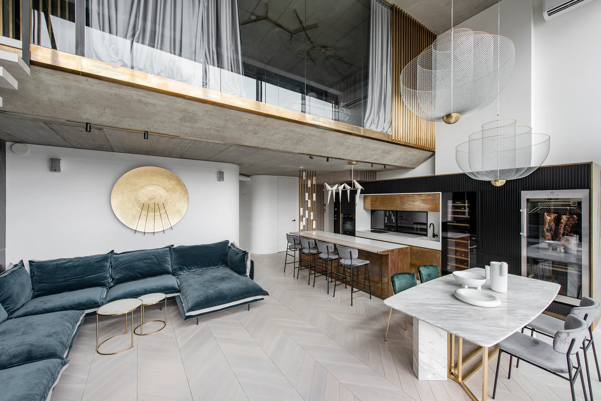 An open-plan loft featuring a teal velvet sectional sofa, a marble dining table, and a concrete mezzanine level with a glass railing.