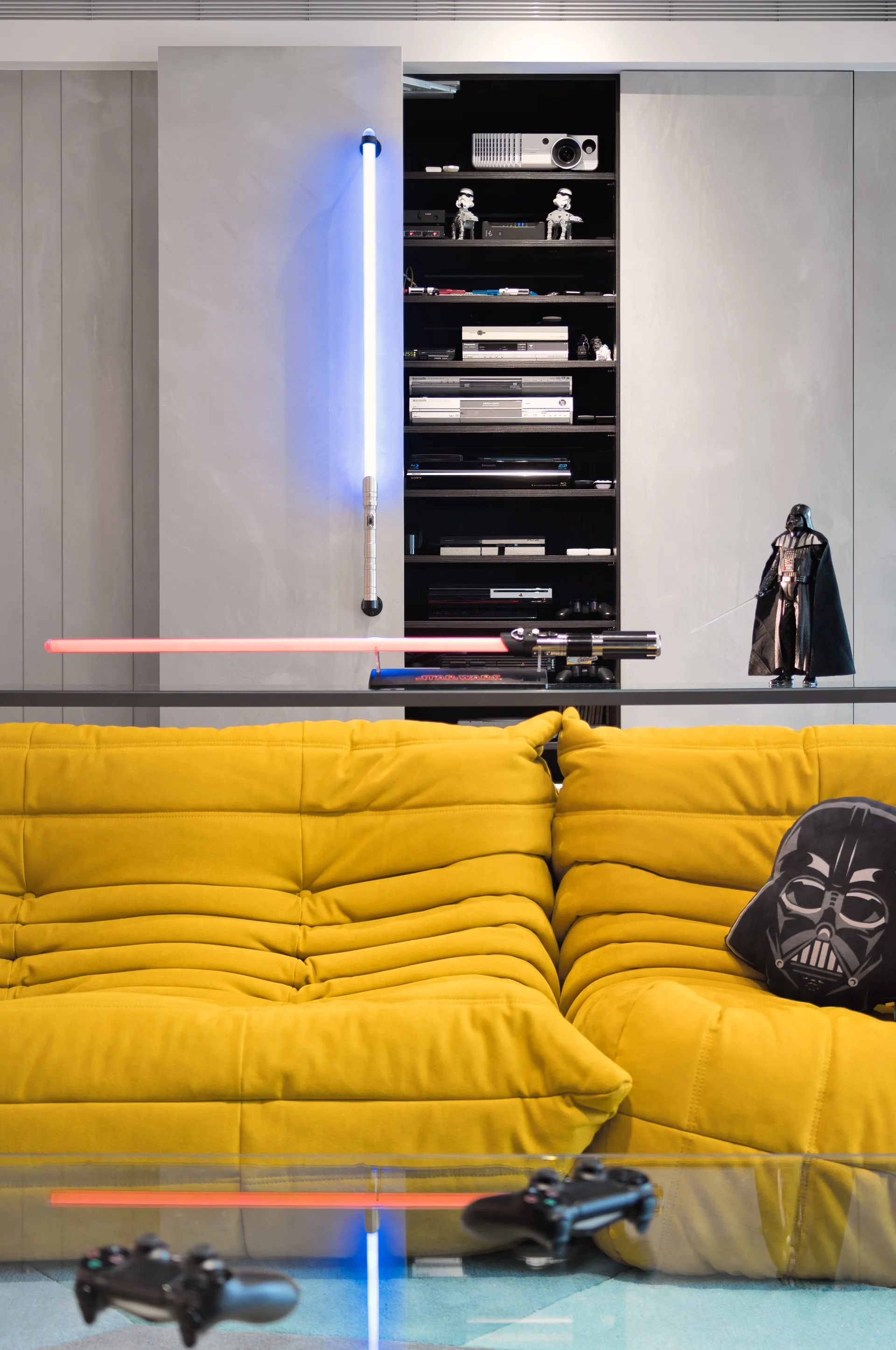 A vibrant yellow tufted sofa sits in front of smooth grey wall panels with a recessed black shelf displaying gaming gear and themed figurines.