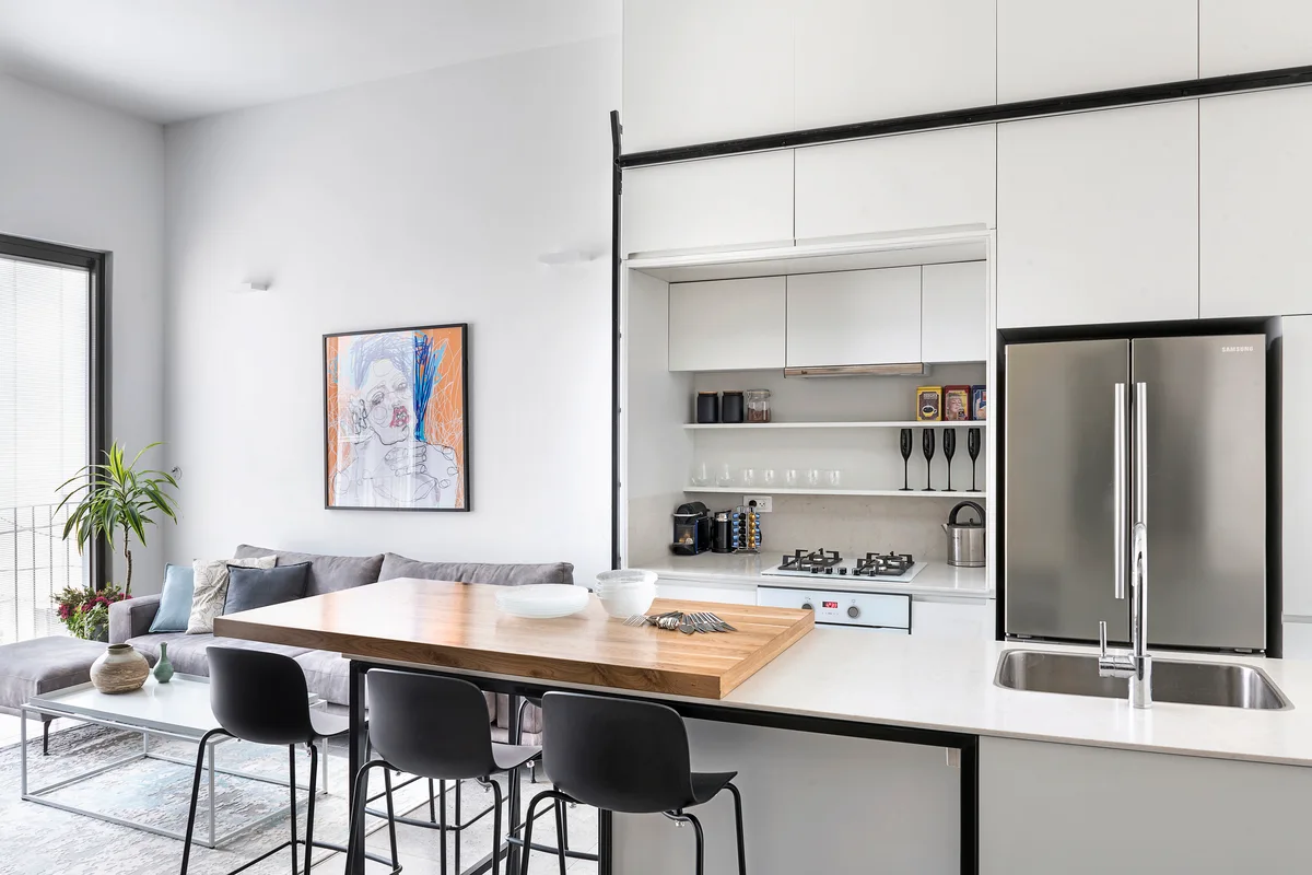A modern kitchen features a white island with an attached wood bar top, black stools, and full-height white cabinetry opening to a living area.